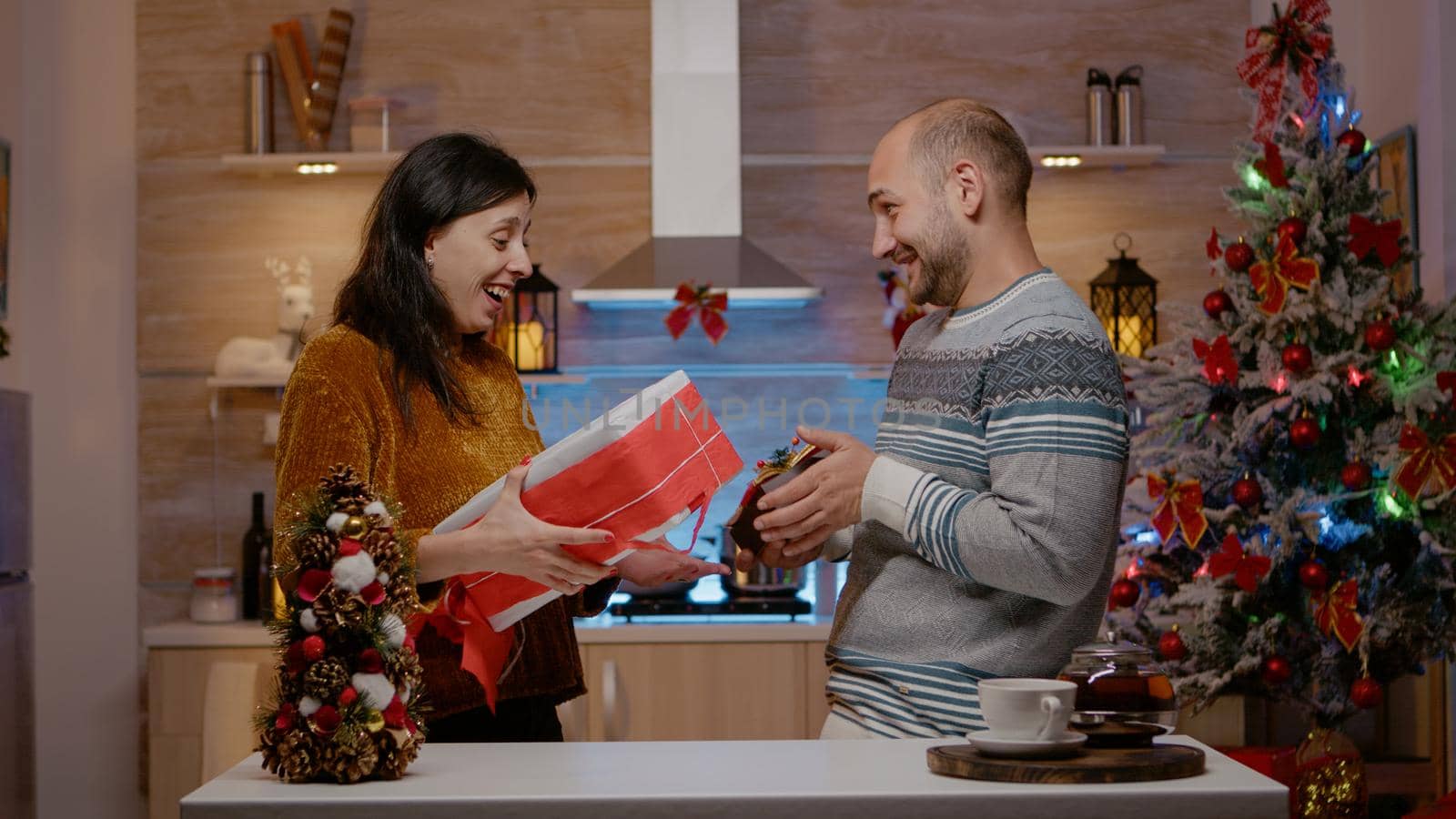 Cheerful couple exchanging presents on christmas eve day for seasonal celebration. Festive people giving gifts to each other celebrating holiday festivity in decorated kitchen.
