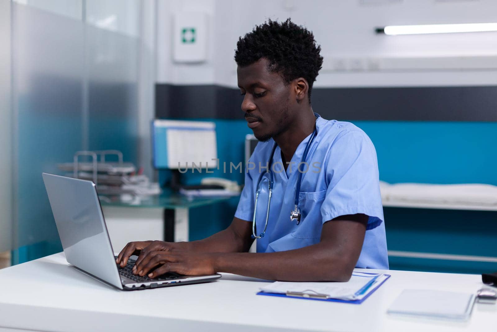 Black young man working as nurse at medical clinic wearing uniform and stethoscope. African american person sitting at desk while using modern laptop, typing on keyboard in cabinet