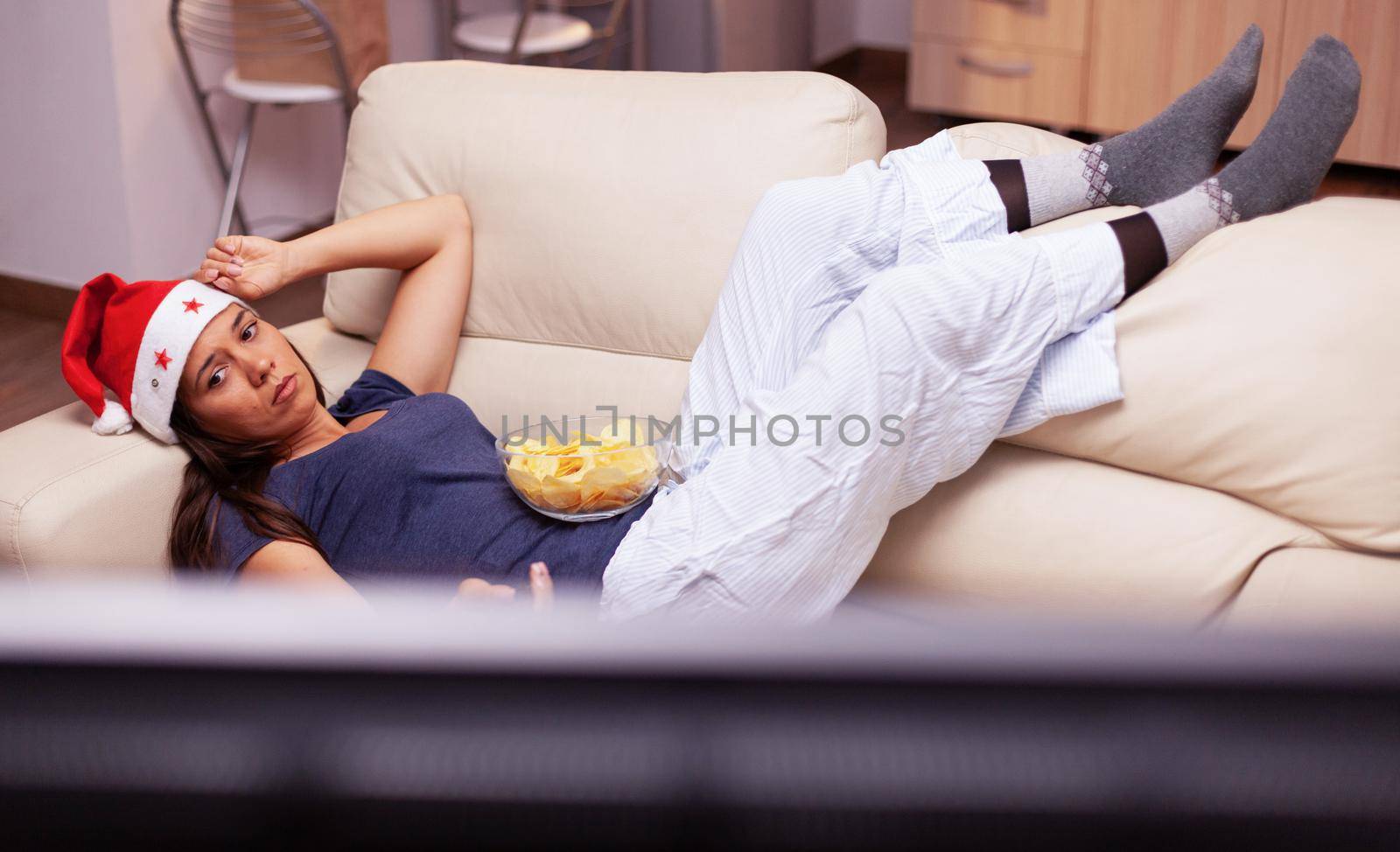 Adult person relaxing on sofa changing channel using remote searching xmas movie on television during christmastime. Woman enjoying winter holiday celebrating christmas season at home