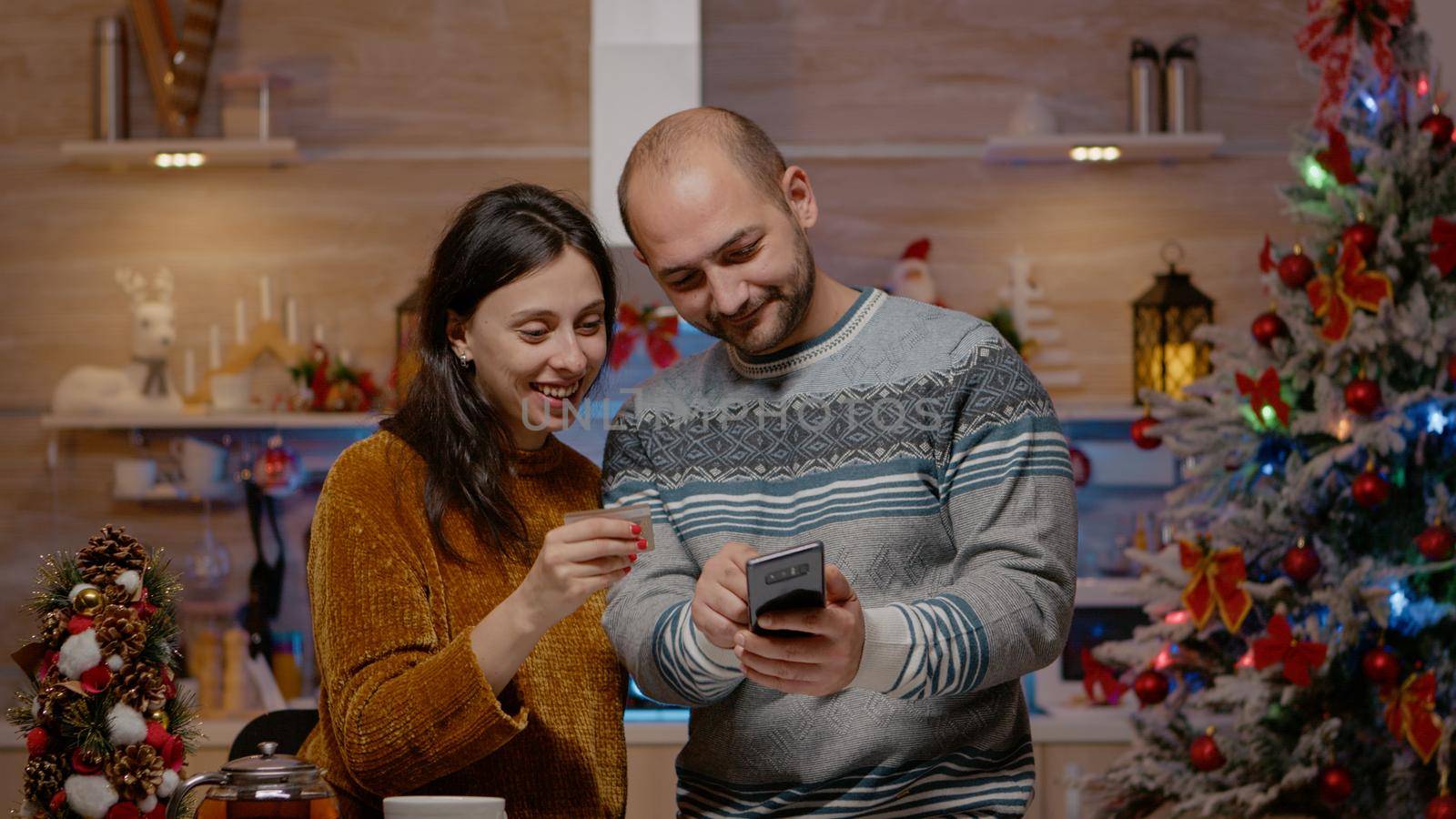 Festive couple buying presents with credit card on smartphone, doing online shopping for christmas eve celebration. People using device paying for gifts in seasonal decorated kitchen