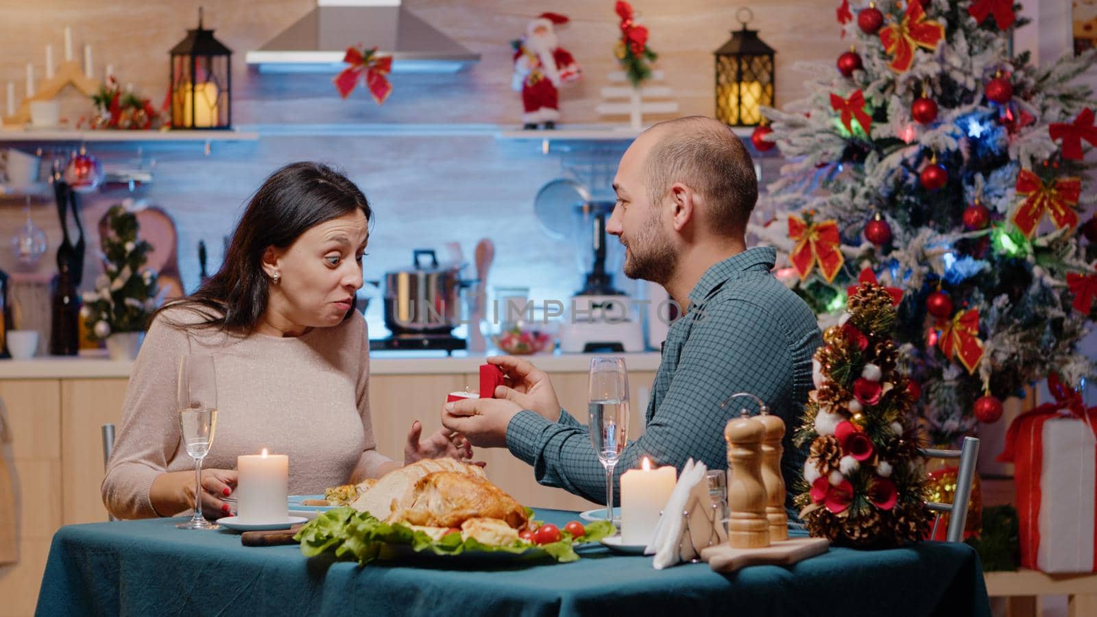 Loving couple enjoying festive dinner with traditional food in decorated kitchen. Man with engagement ring proposing to woman on christmas eve feeling happy. Romantic holiday celebration