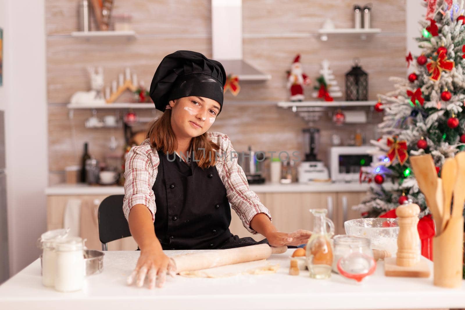 Portrait of kid wearing apron preparing homemade dough using rolling pin enjoying winter season in xmas decorated culinary kitchen. Child cooking gingerbread dessert celebrating christmas holiday