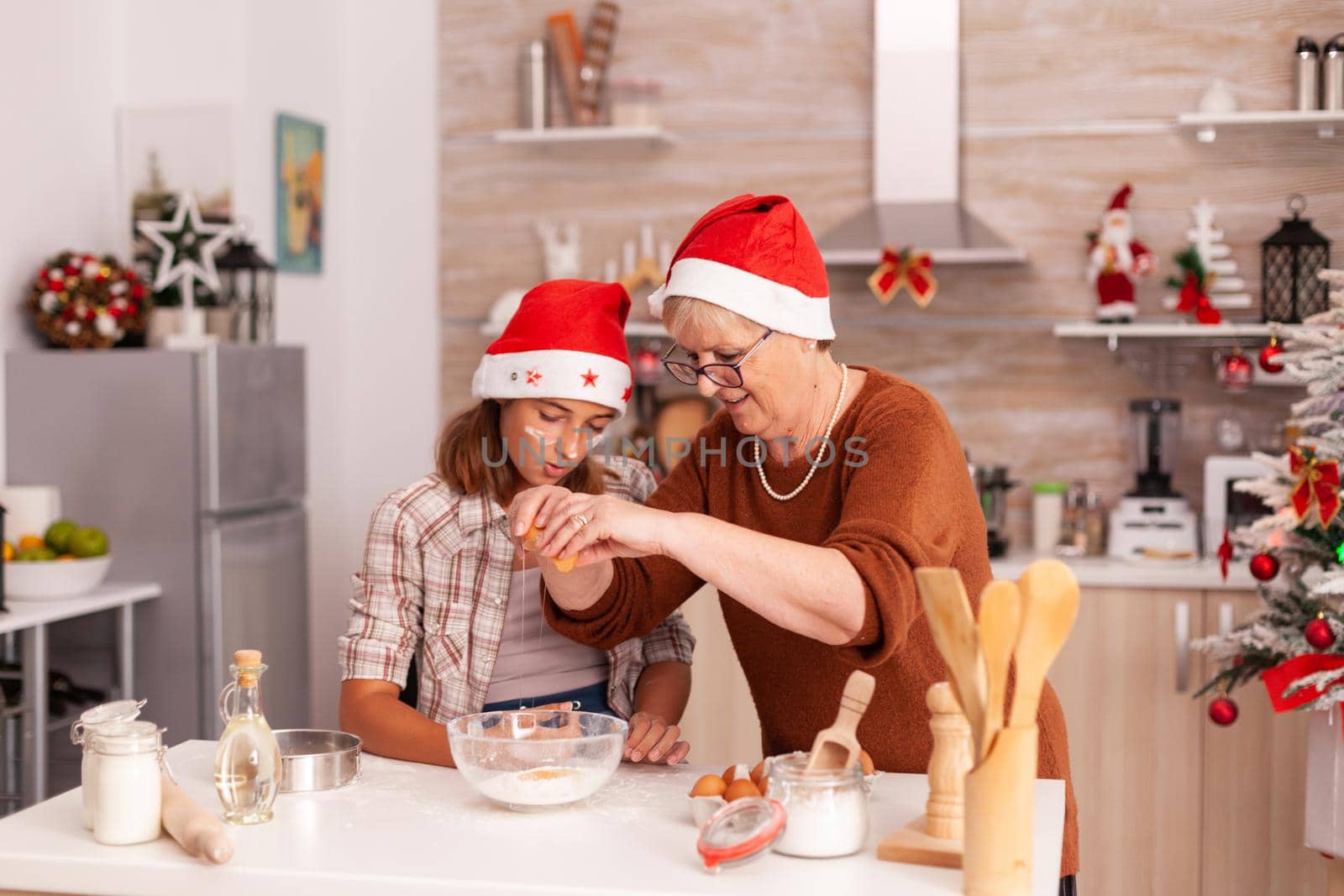 Grandmother showing to children how to prepare cookies dough breaking eggs in ingredients bowl enjoying winter holiday together in xmas decorated kitchen. Family celebrating christmas season