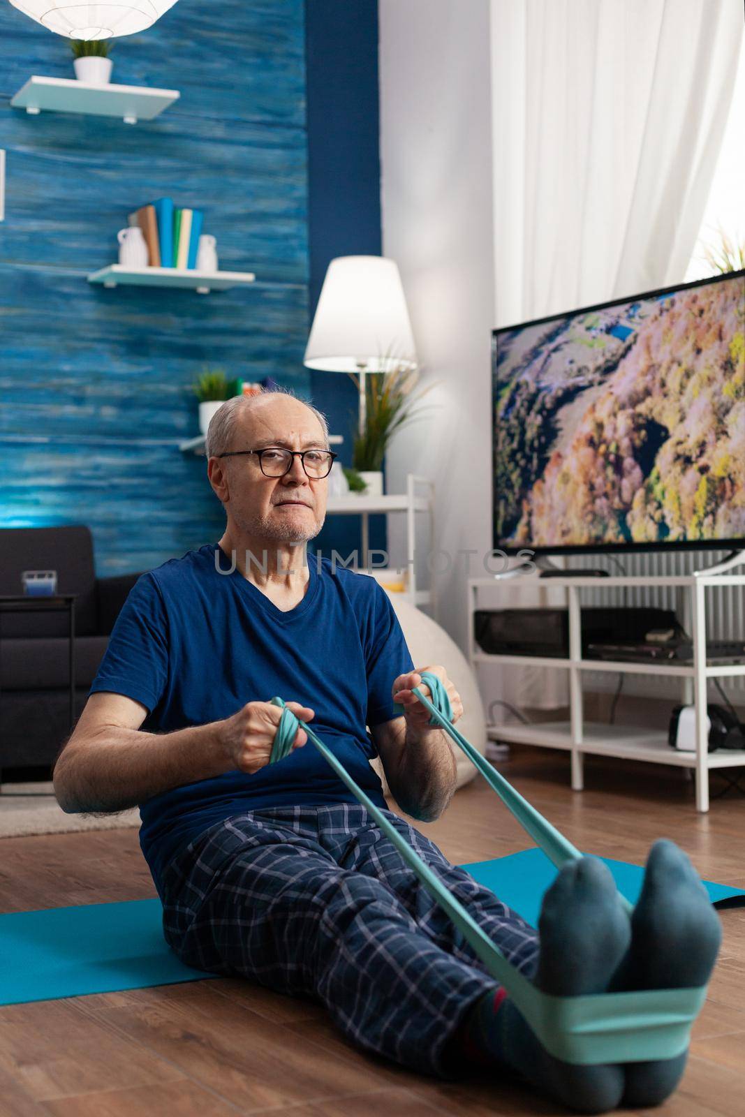 Focused pensioner sitting on yoga mat stretching legs muscle using fitness elastic band by DCStudio