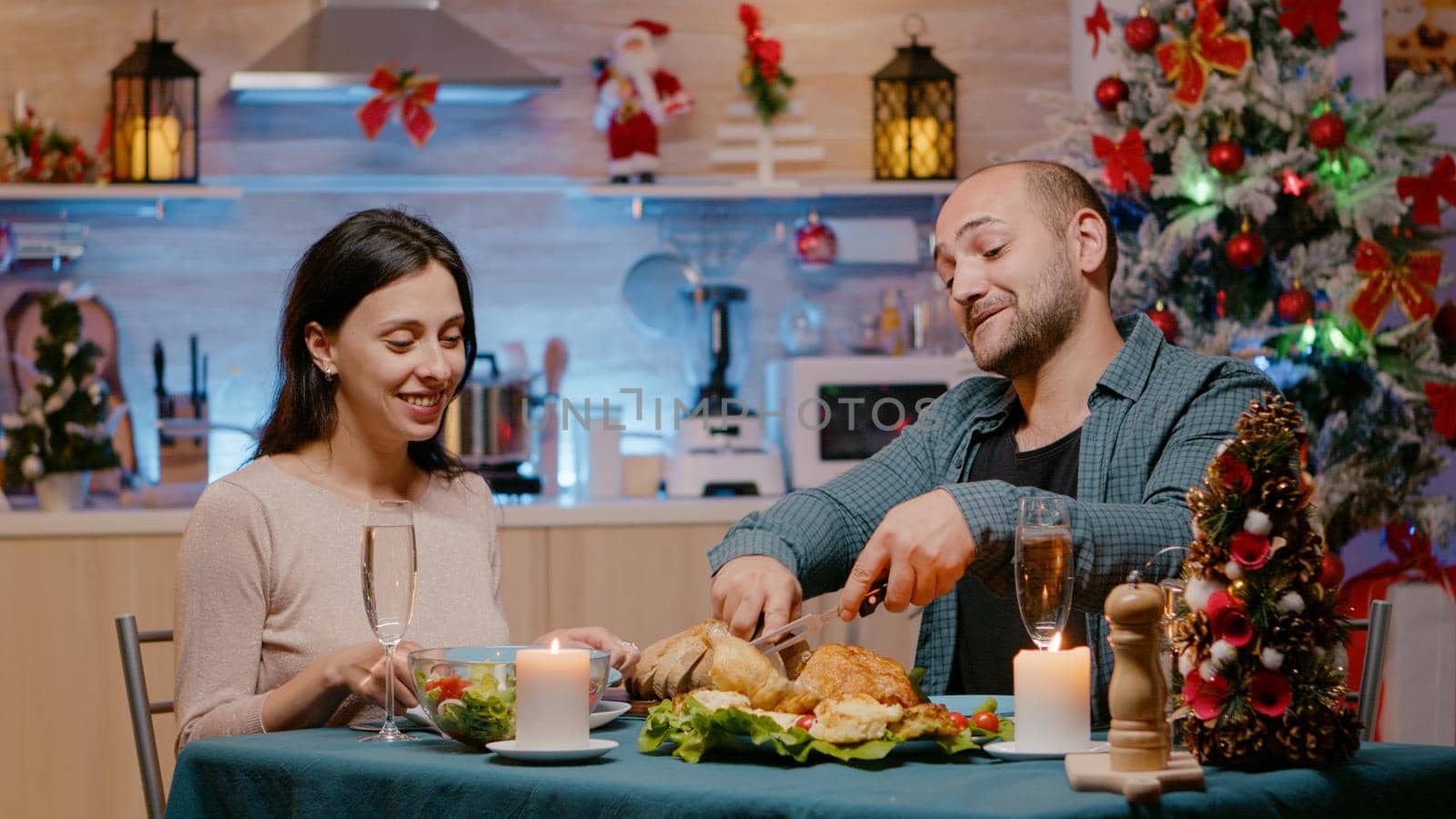 Cheerful couple celebrating christmas eve with festive dinner in decorated kitchen. Man and woman eating seasonal meal and drinking champagne while having candles on table for celebration.