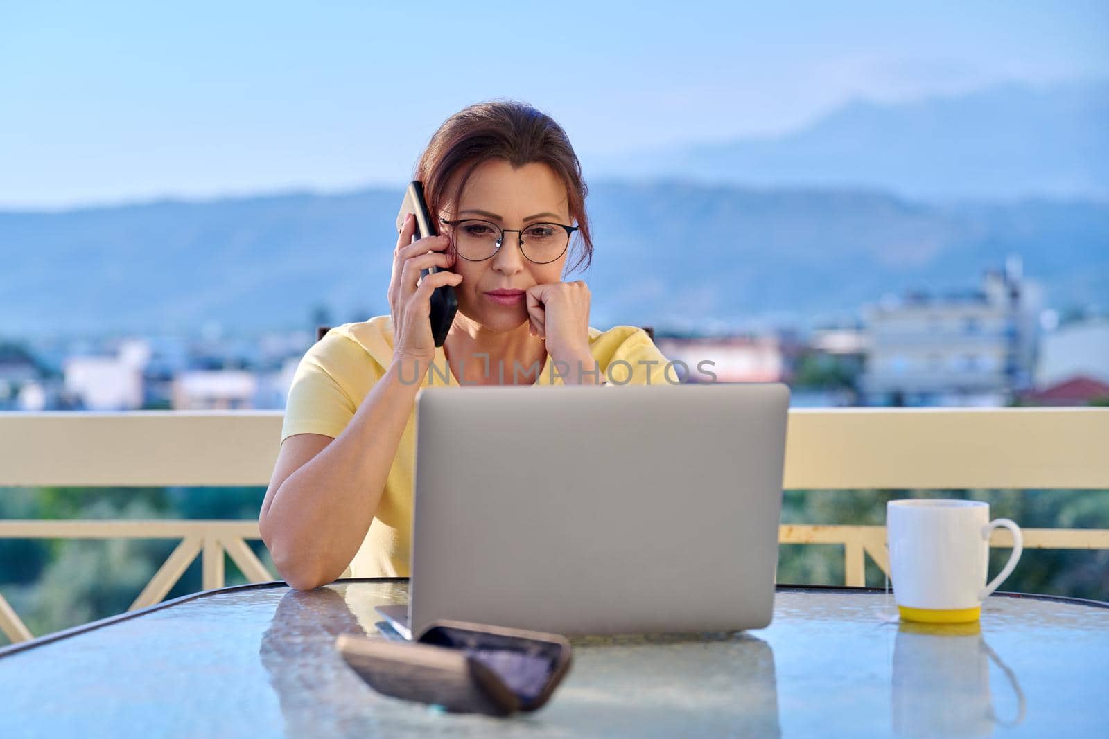 Serious middle aged woman with laptop smartphone on outdoor balcony. Concentrated 40s mature business woman working remotely. Freelance, technology, work from home office, online training, blog, vlog