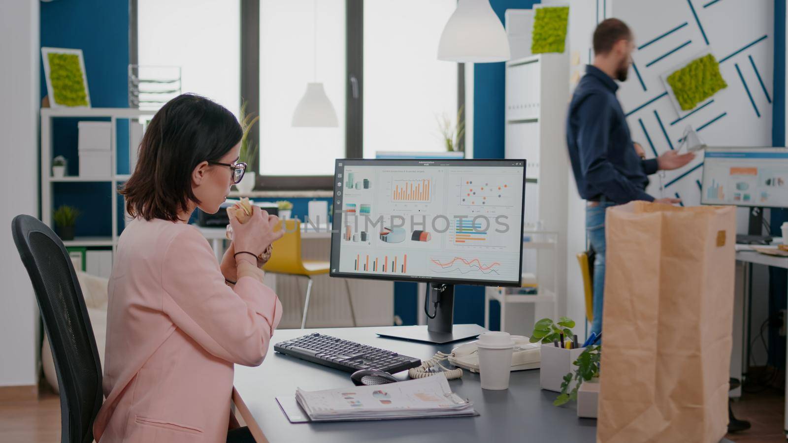 Executive manager eating tasty sandwich while analyzing marketing strategy at desk in workplace. Takeout order food delivery in corporate place, lunch meal break package delivered at startup office.