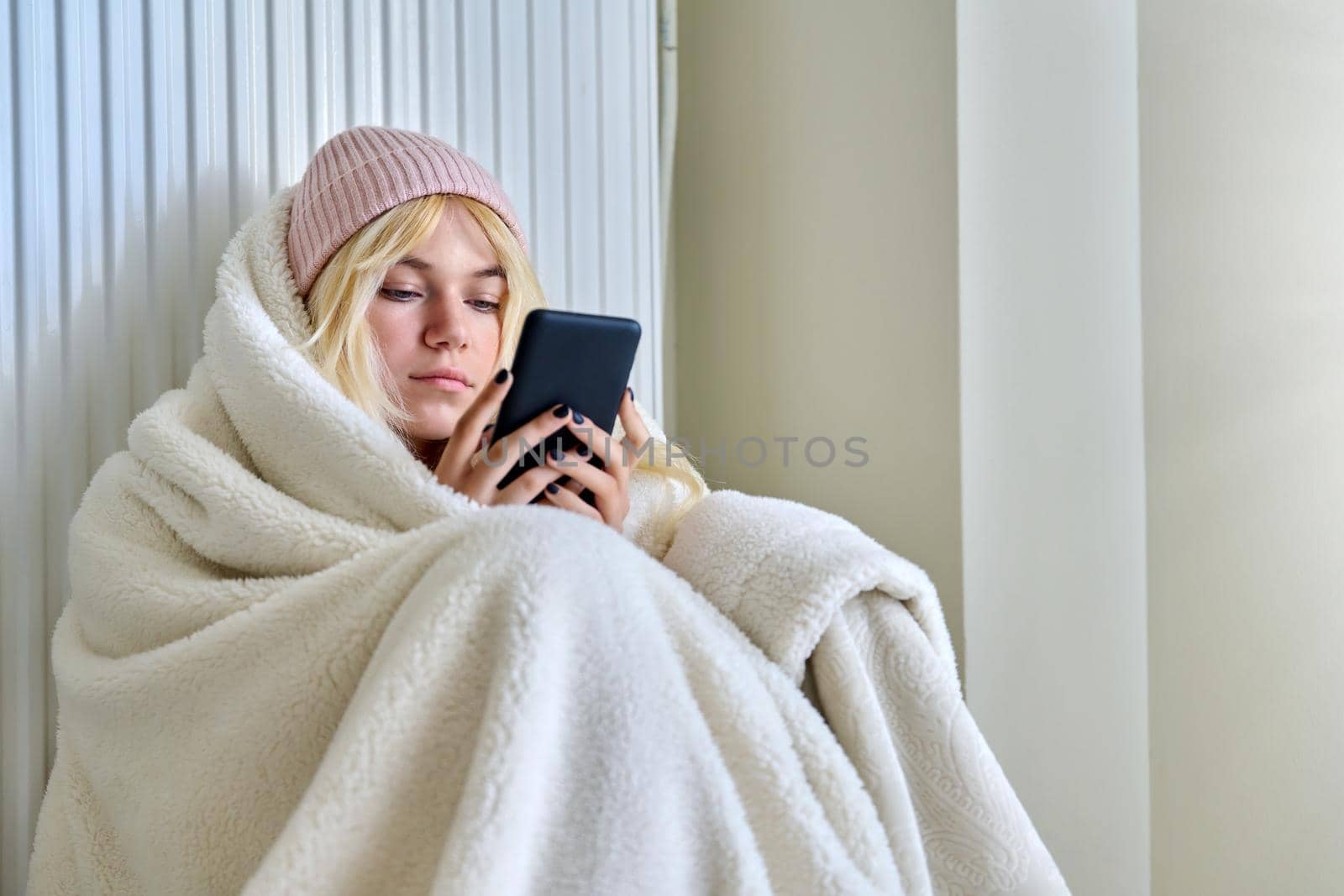 The cold season, cold winter and autumn, a frozen female teenager in a hat under a blanket sitting near a heating radiator with a smartphone in her hands