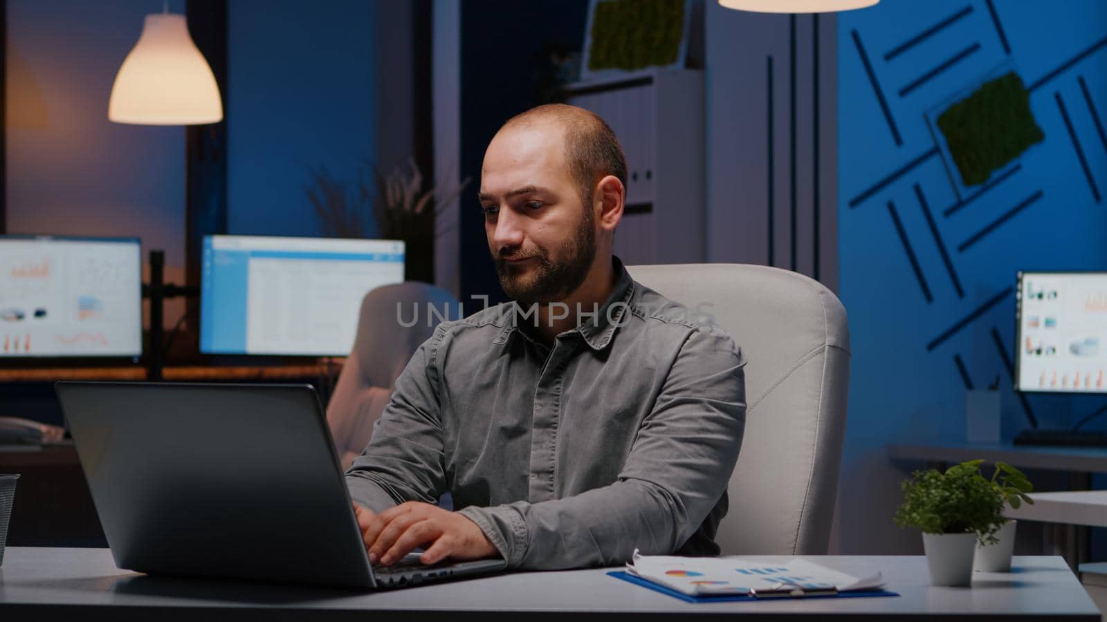 Overworked businessman typing financial statistics on laptop sitting at desk table in startup company office late at night. Workaholic entrepreneur planning economic project before company deadline