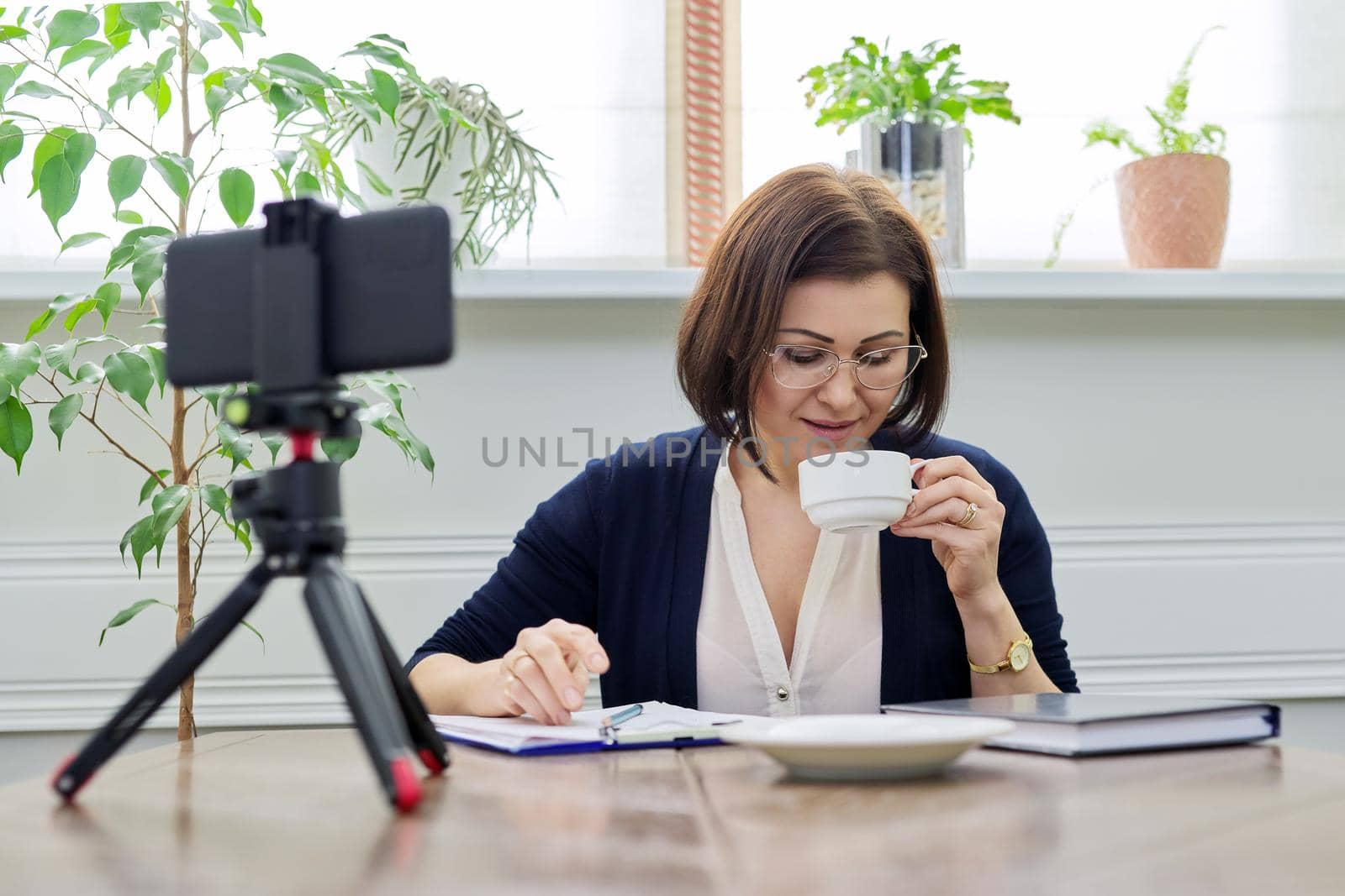 Middle aged business woman, counselor, mentor in video meeting, using smartphone on tripod. Positive female with cup of coffee talking looking in webcam. Video consultations, freelancing, remote business, online education