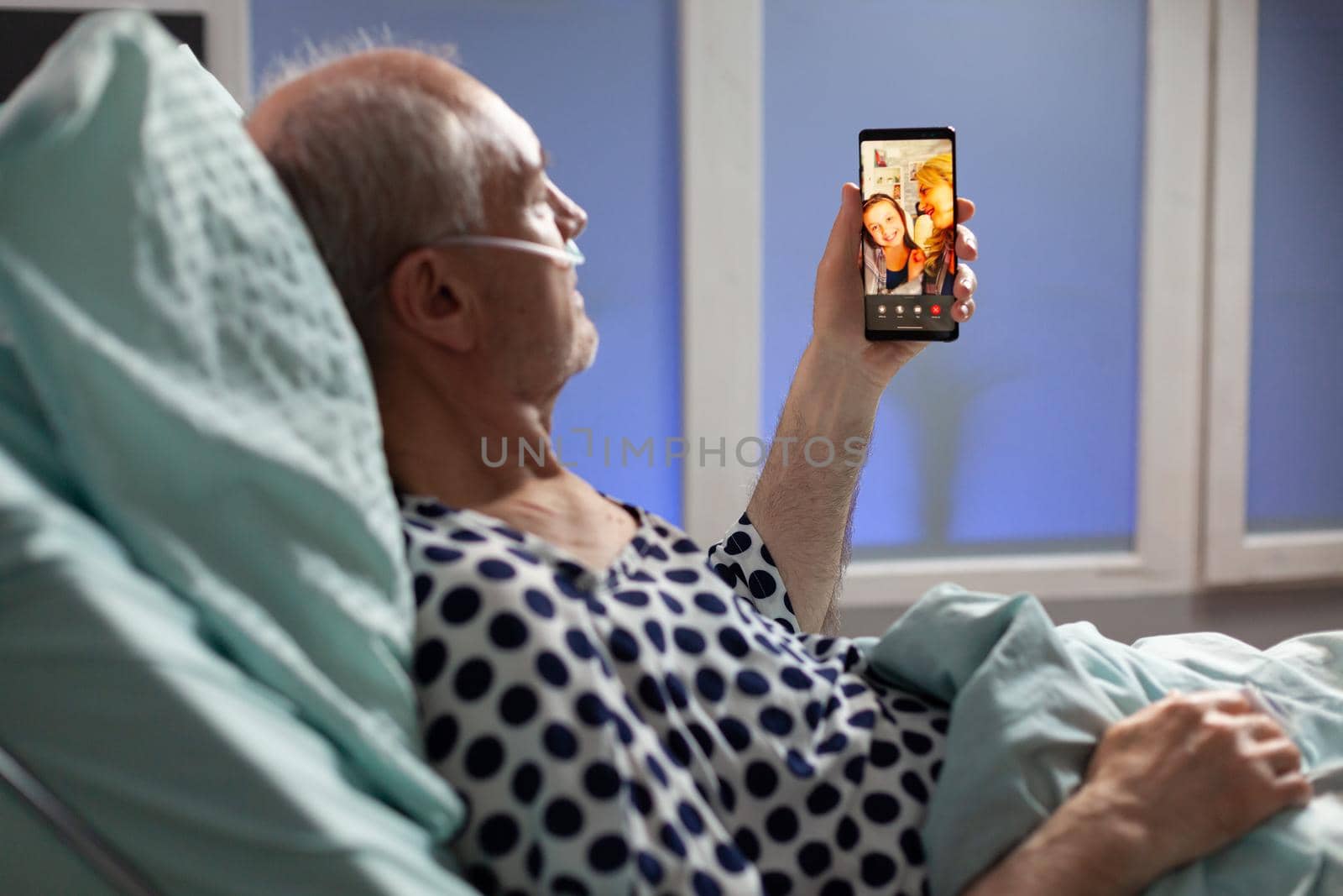 Senior sick man breathing through oxygen tube saying hello to relatives, laying in hospital bed holding phones, discussing about recovery. Old man waiting for treatment.