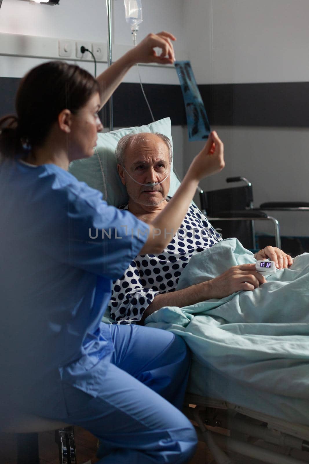 Old patient with lungs illness breathing using oxygen mask laying in hospital bed, listening nurse showing x-ray explayning diagnosis before surgery.