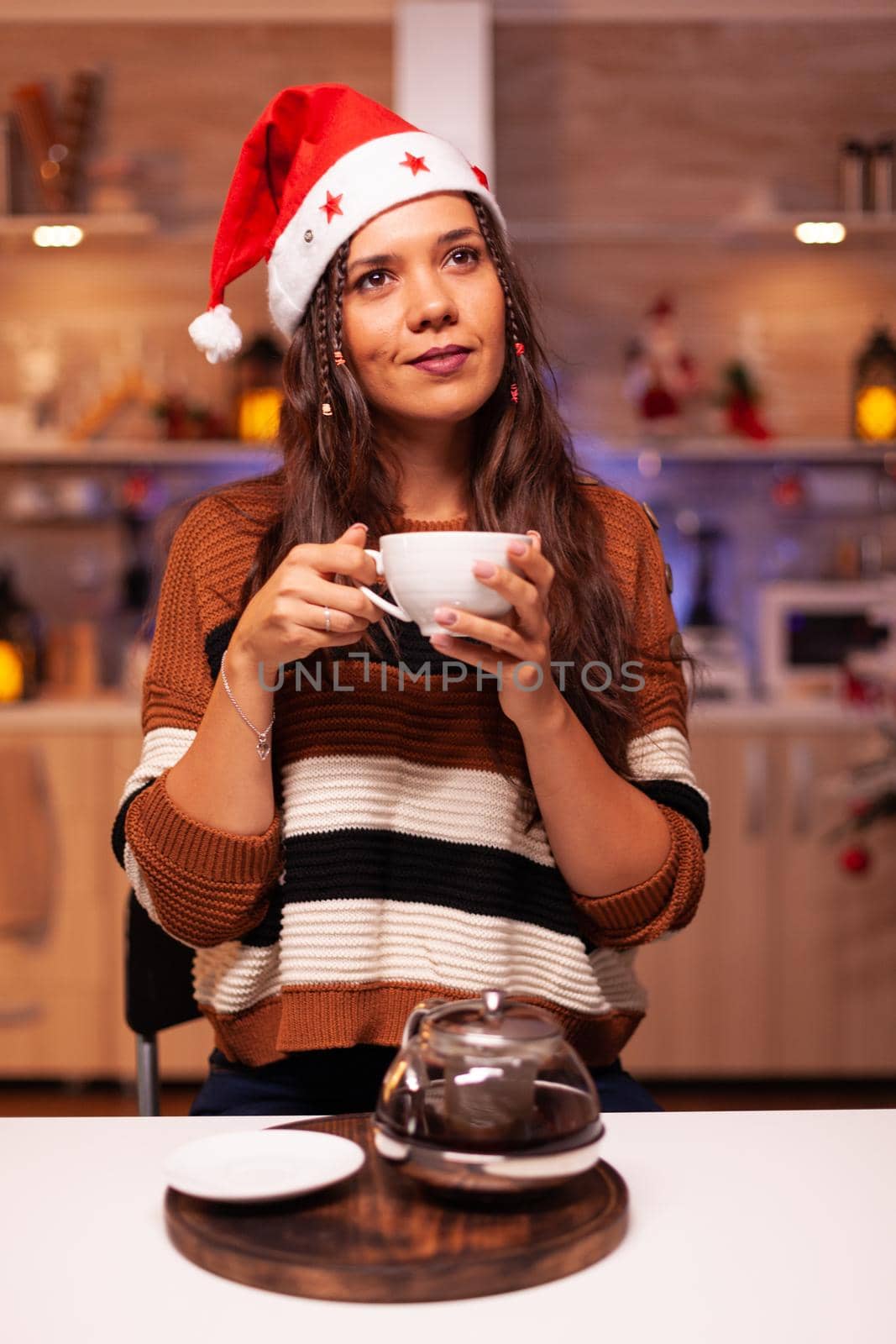 Portrait of caucasian woman with santa hat smiling in kitchen decorated with ornaments, lights and tree for christmas season. Cheerful adult thinking about winter celebration dinner