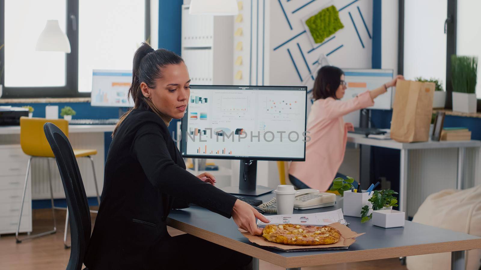 Businesswoman having lunchtime eating takeaway delivery pizza slice at desk in company office working at financial graphs. Takeout lunch meal break package delivered at startup business corporate.