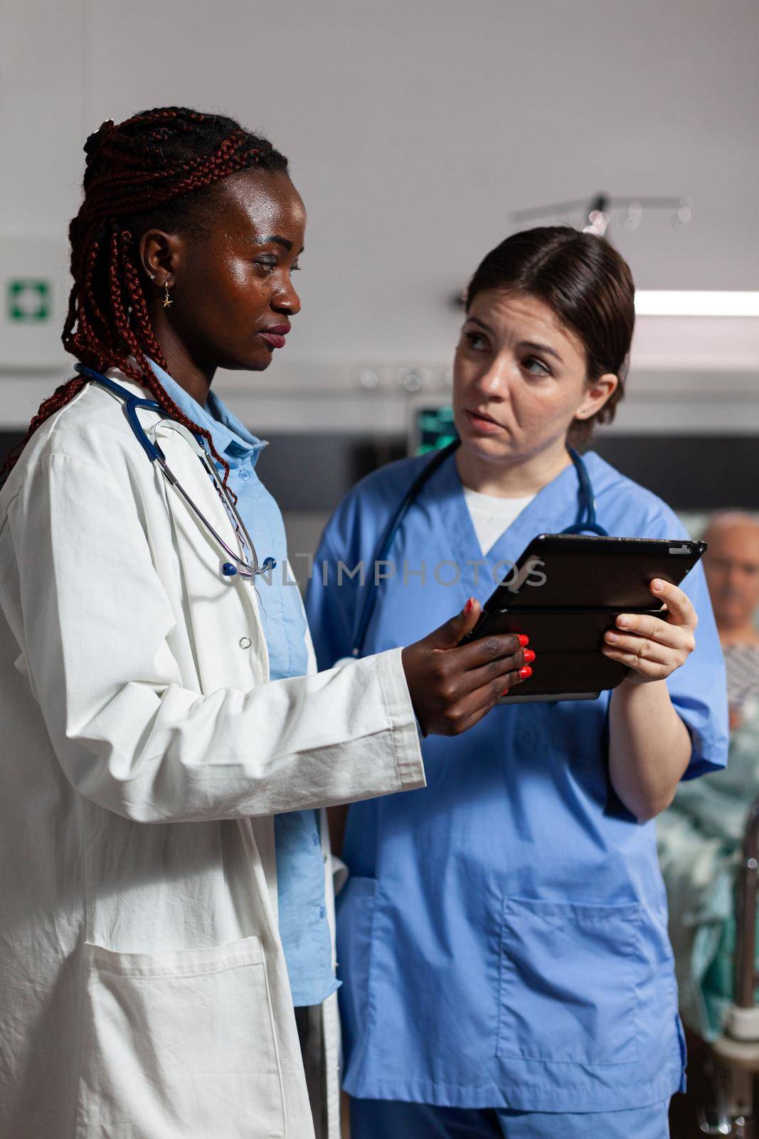 Medical nurse asking advice from african american doctor, holding tablet pc in hospital room, about treatment for sick, ill, unwell senior man laying in bed.
