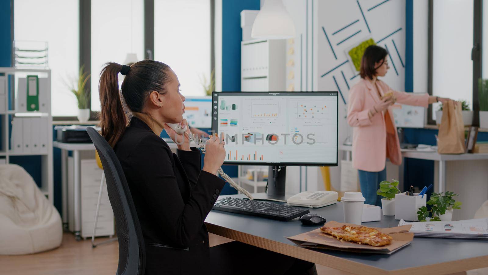 Multitasking businesswoman talking at landline explaining company graphs to remote collegue while having takeaway delivery meal food order at desk. Manager eating tasting pizza slice