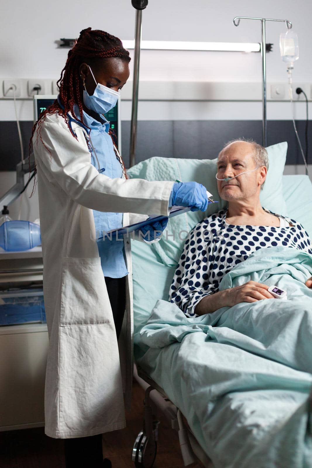 African american doctor with surgical mask reading diagnosis from clipboard to sick ill unwell senior patient laying in bed, breathing with help from oxygen mask, listening discussing with medical staff about recovery.