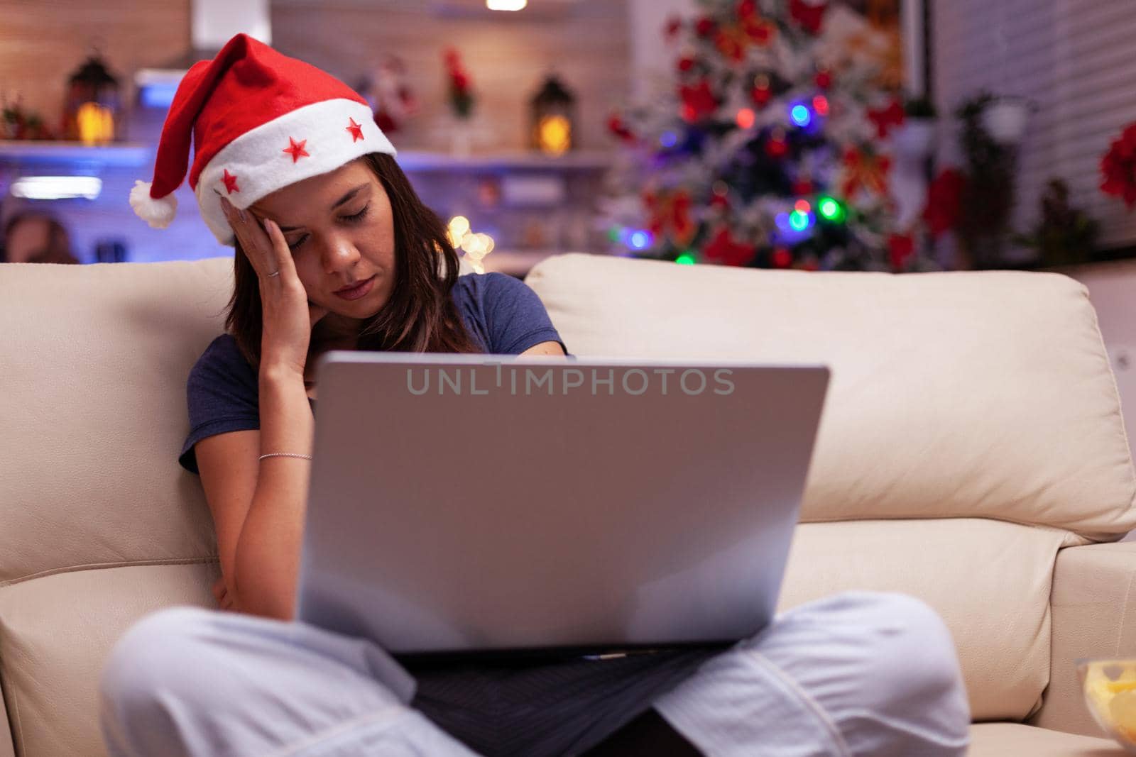 Tired exhausted woman falling asleep while reading business email on laptop computer sitting on sofa in xmas decorated kitchen. Girl enjoying winter holiday celebrating christmas season