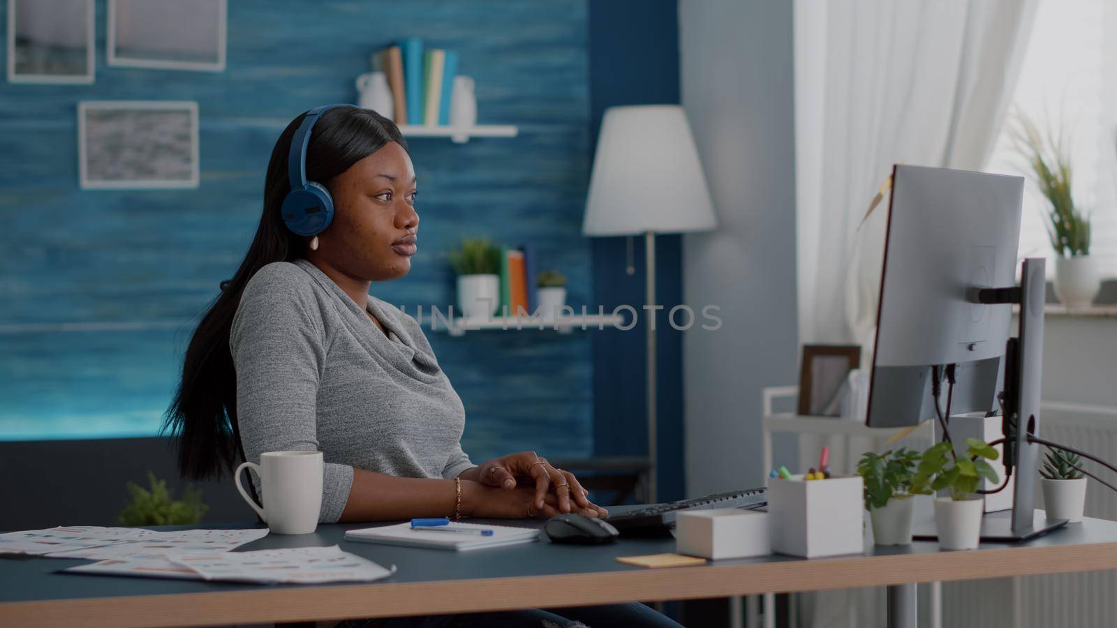 Student with black skin having headphone puts listening online univeristy course using elearning platform sitting at desk in living room. African american woman working remote from home