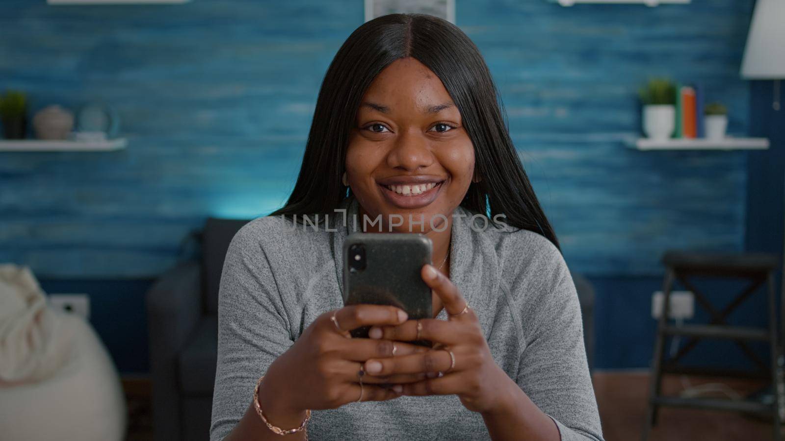 Black student looking into camera while browsing social media typing message socializing with friends on modern smartphone. Young woman sitting at desk in living room sharing group conversation