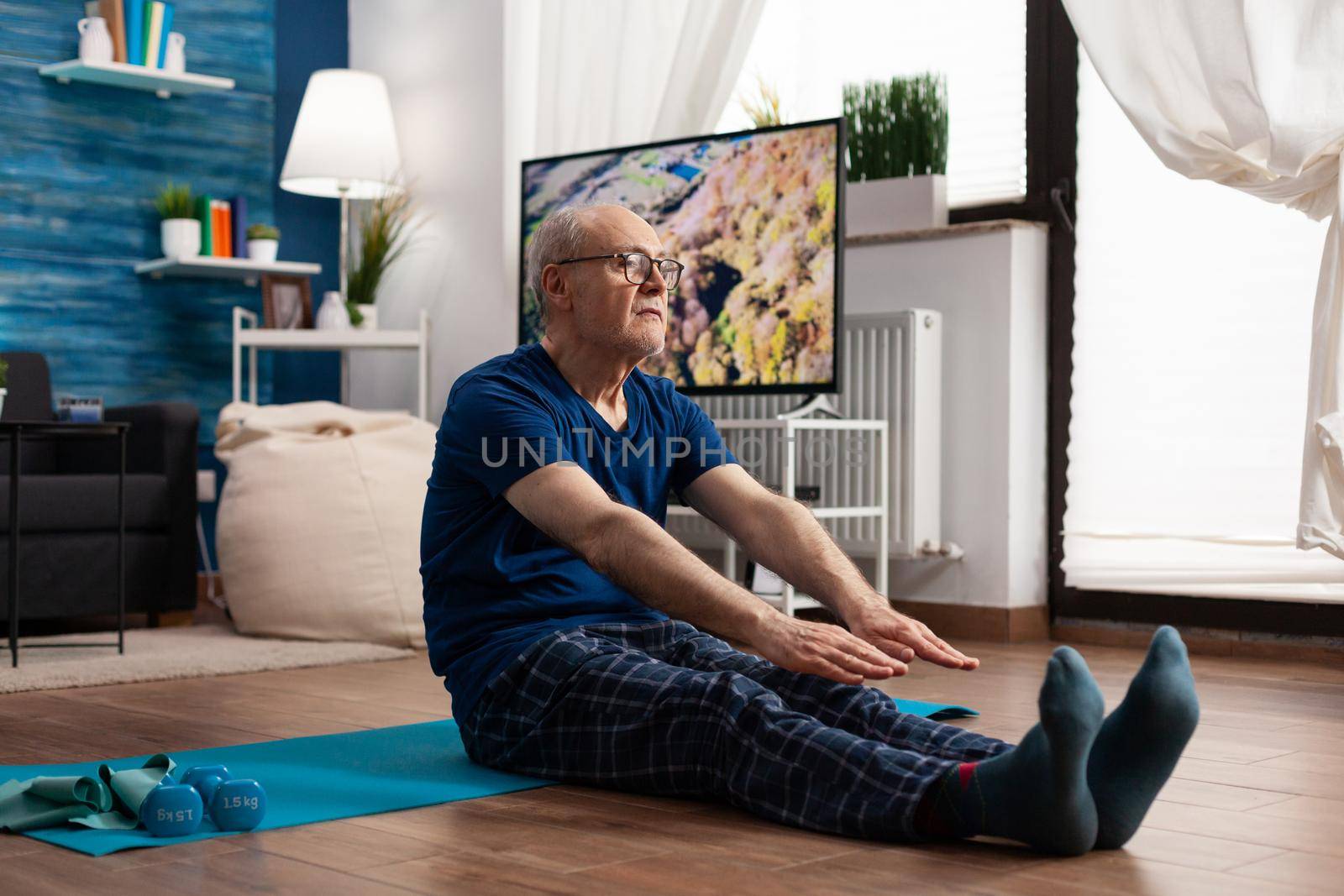 Focused pensioner wearing sportswear sitting on yoga mat stretching legs muscle during aerobics workout exercising muscles resistance. Senior man doing body exercise slimming weight