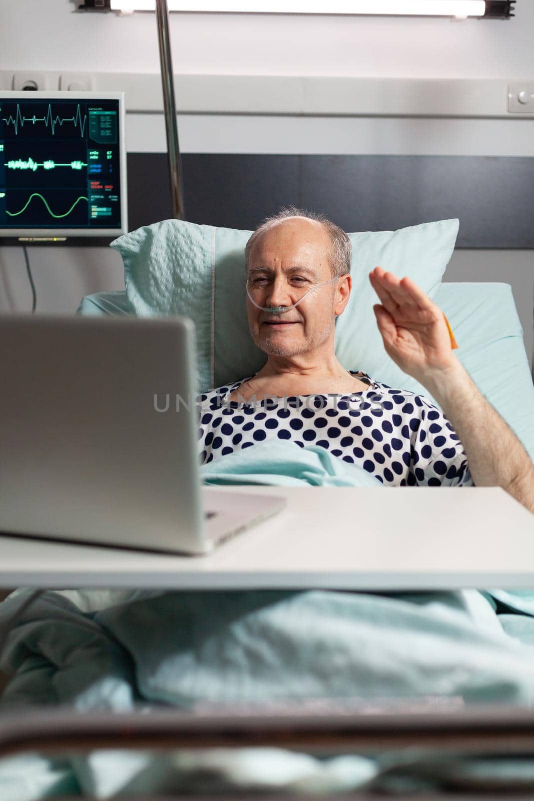 Cheerful sick senior man waving at camera during video conference using laptop laying in bed, breathing through oxygen tube. Man recovering after surgery and treatment.