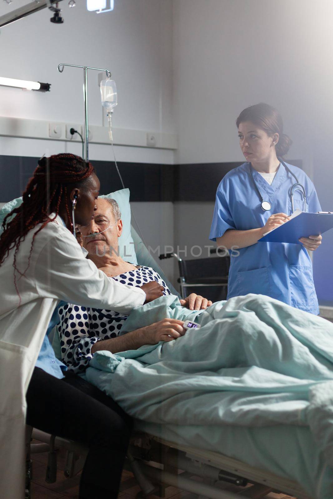 Cardiologist in uniform examining senior man during medical visit, listening heart using stehoscope to listen heart. Man with sick lungs breathing through oxygen tube and oximeter attached on finger.