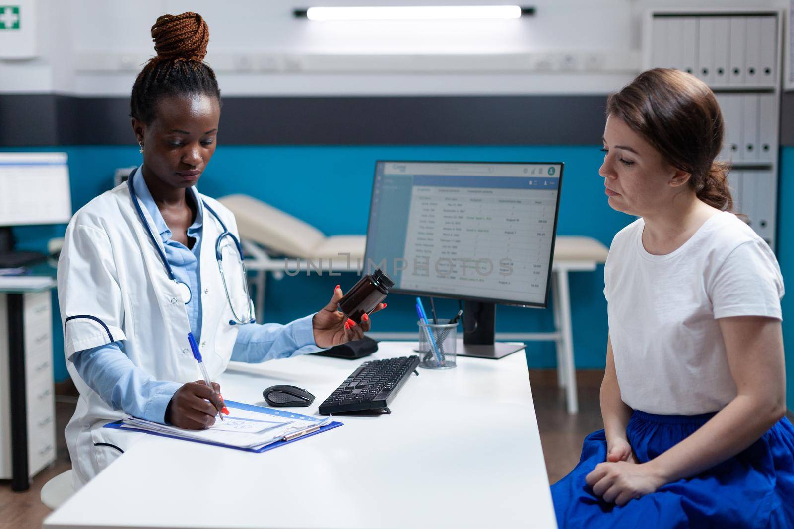 African american therapist doctor holding pills bottle explaining disease symptoms to sick woman patient during clinical appointment in hospital office. Practitioner medic writing medication treatment