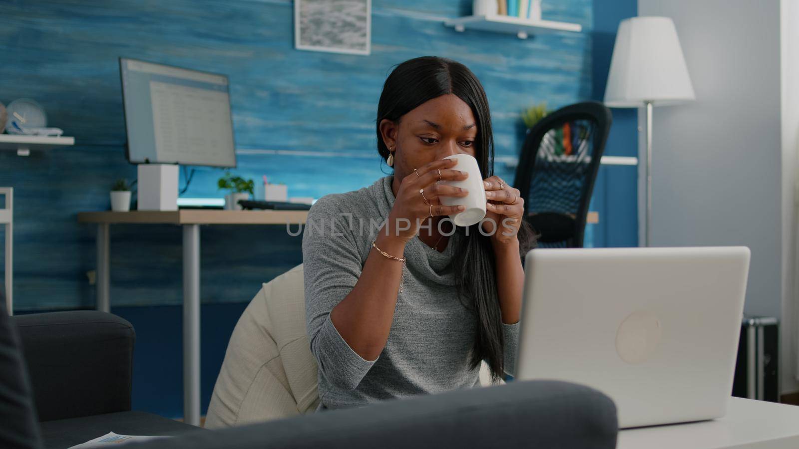 Black student smiling while working at social media blog article typing communication project sitting in living room. Blogger woman drinking coffee composing email solving webinar problem