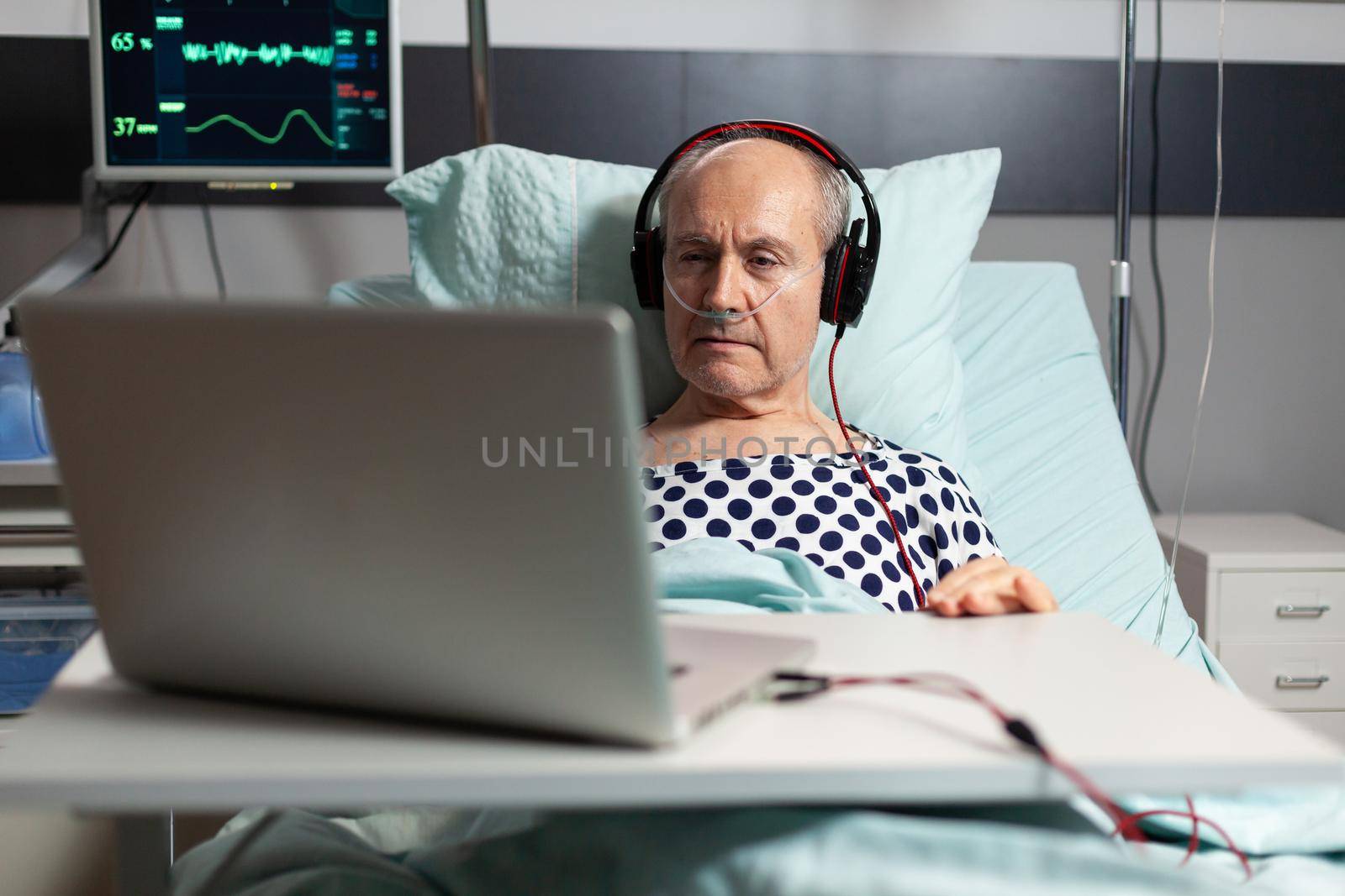 Senior sick man in hospital bed breathing through oxygen mask, listening music on headphones, looking at laptop. Old man monitored by modern equipment.