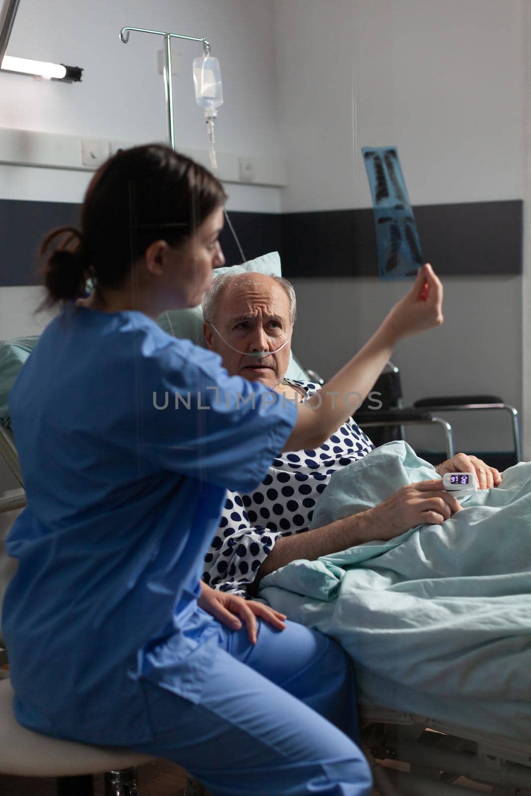 Old patient with lungs illness breathing using oxygen mask laying in hospital bed, listening nurse showing x-ray explayning diagnosis before surgery.