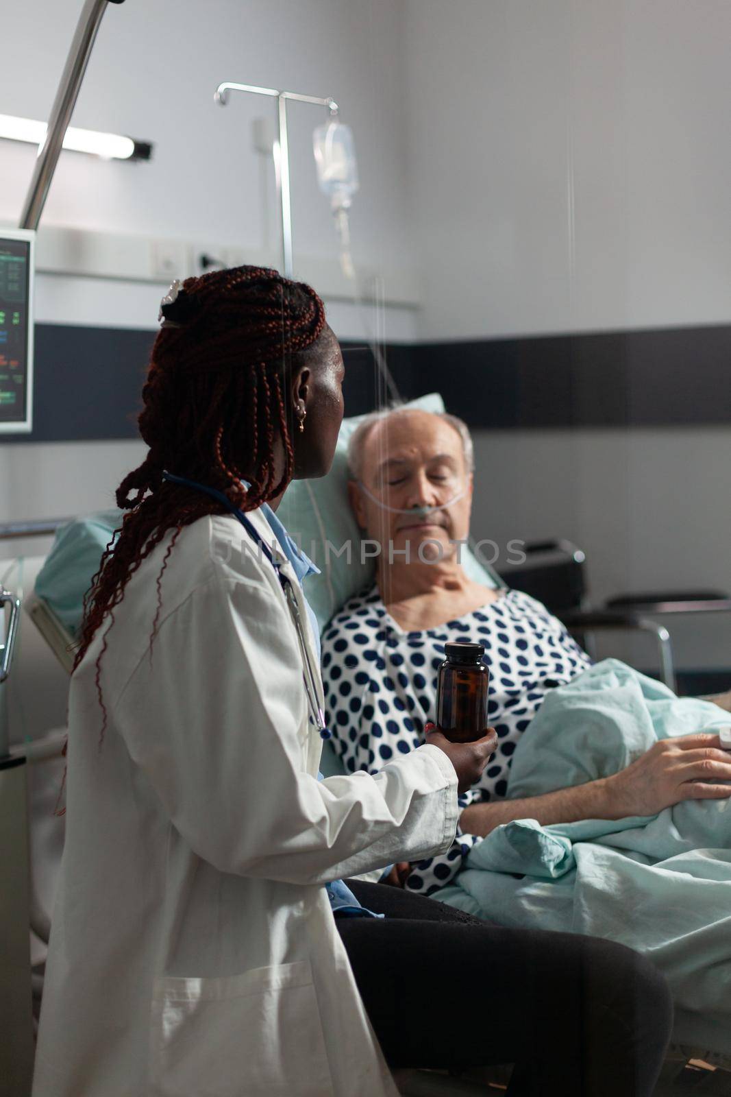 Senior man breathing with help from oxygen tube laying in hospital bed, listening african american doctor explaying diagnosis treatmant holding pills bottle for recovery.