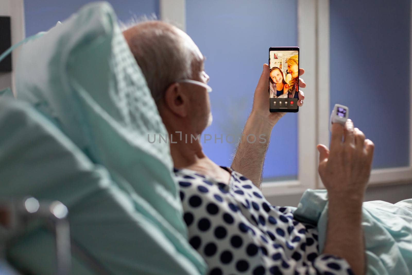 Senior sick man breathing through oxygen tube saying hello to relatives, laying in hospital bed holding phones, discussing about recovery. Old man waiting for treatment.