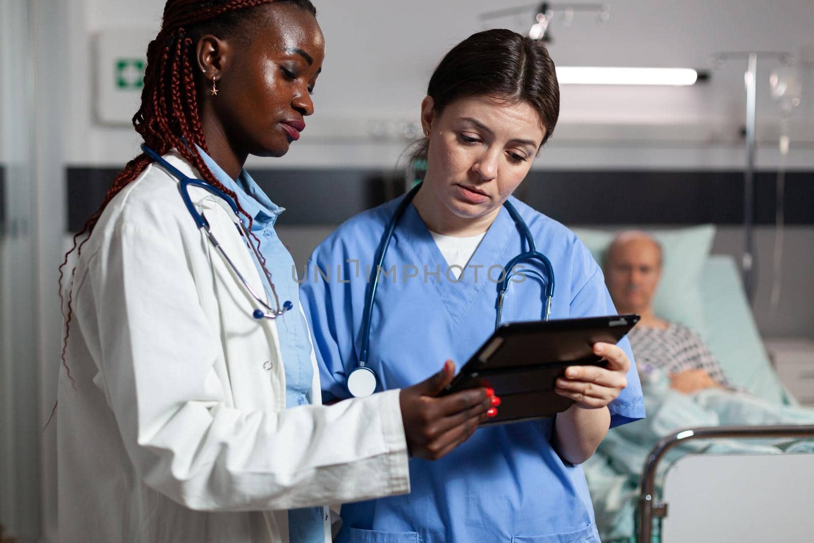 Medical nurse asking advice from african american doctor, holding tablet pc in hospital room, about treatment for sick, ill, unwell senior man laying in bed.