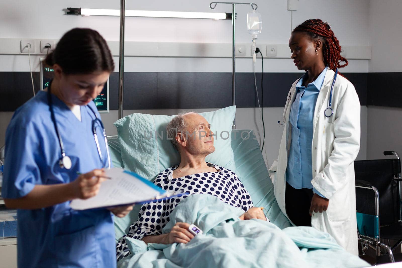 African doctor ward in hospital room discussing with sick senior man laying in bed breathing through oxygen tube with iv dirp bag attached, during consultation. Medical nurse taking notes on clipboard.