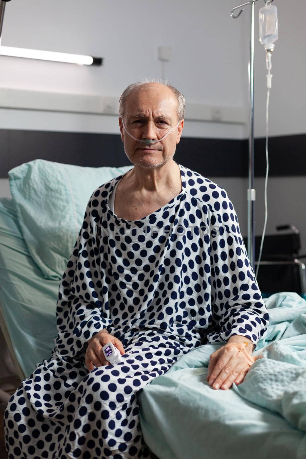 Portrait of sad, unwell senior man sitting on the edge of hospital bed with iv drip attached and breathing with help from oxygen mask, looking at camera.