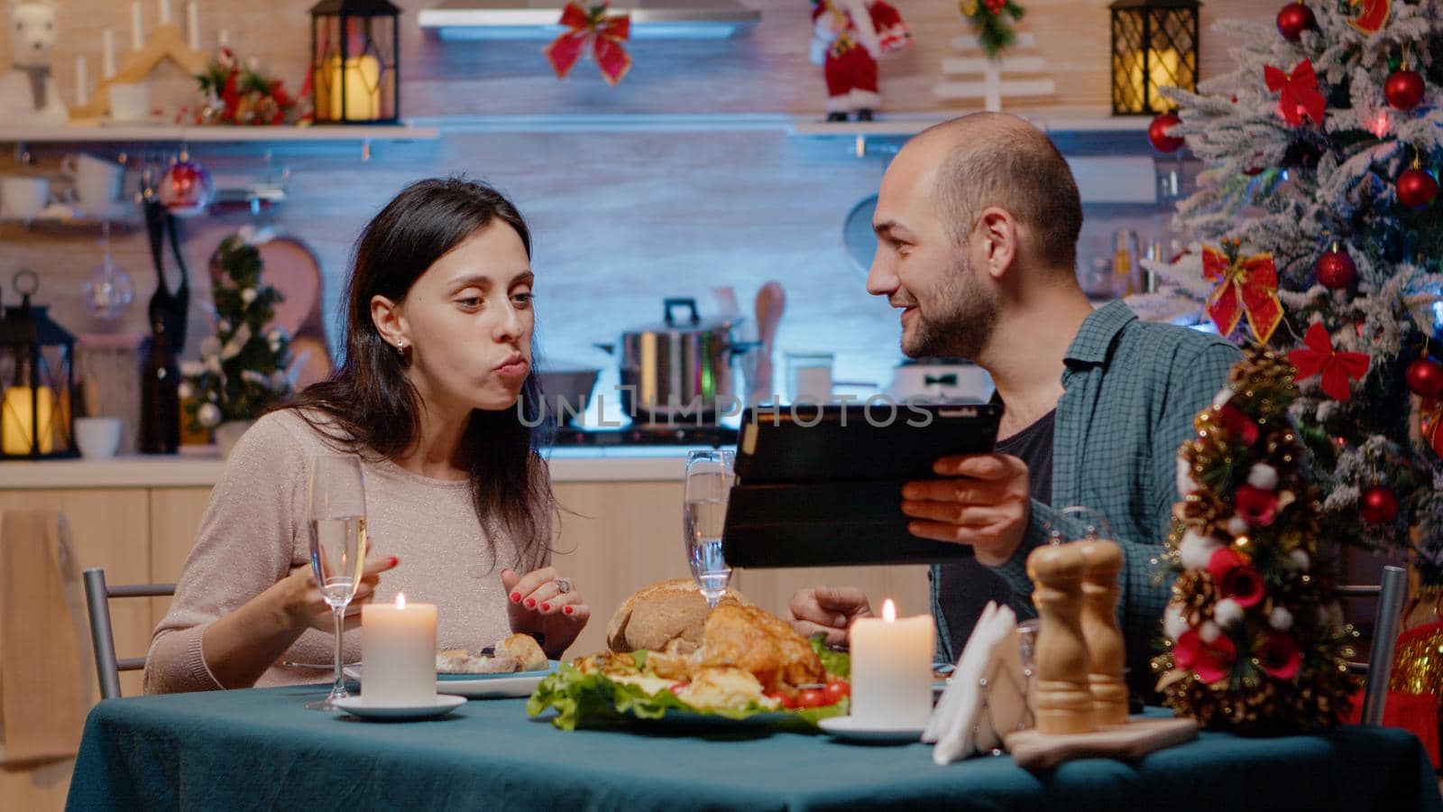 Couple enjoying christmas dinner and looking at tablet laughing and celebrating festive holiday at home. Man and woman watching device while clinking glasses with champagne and eating