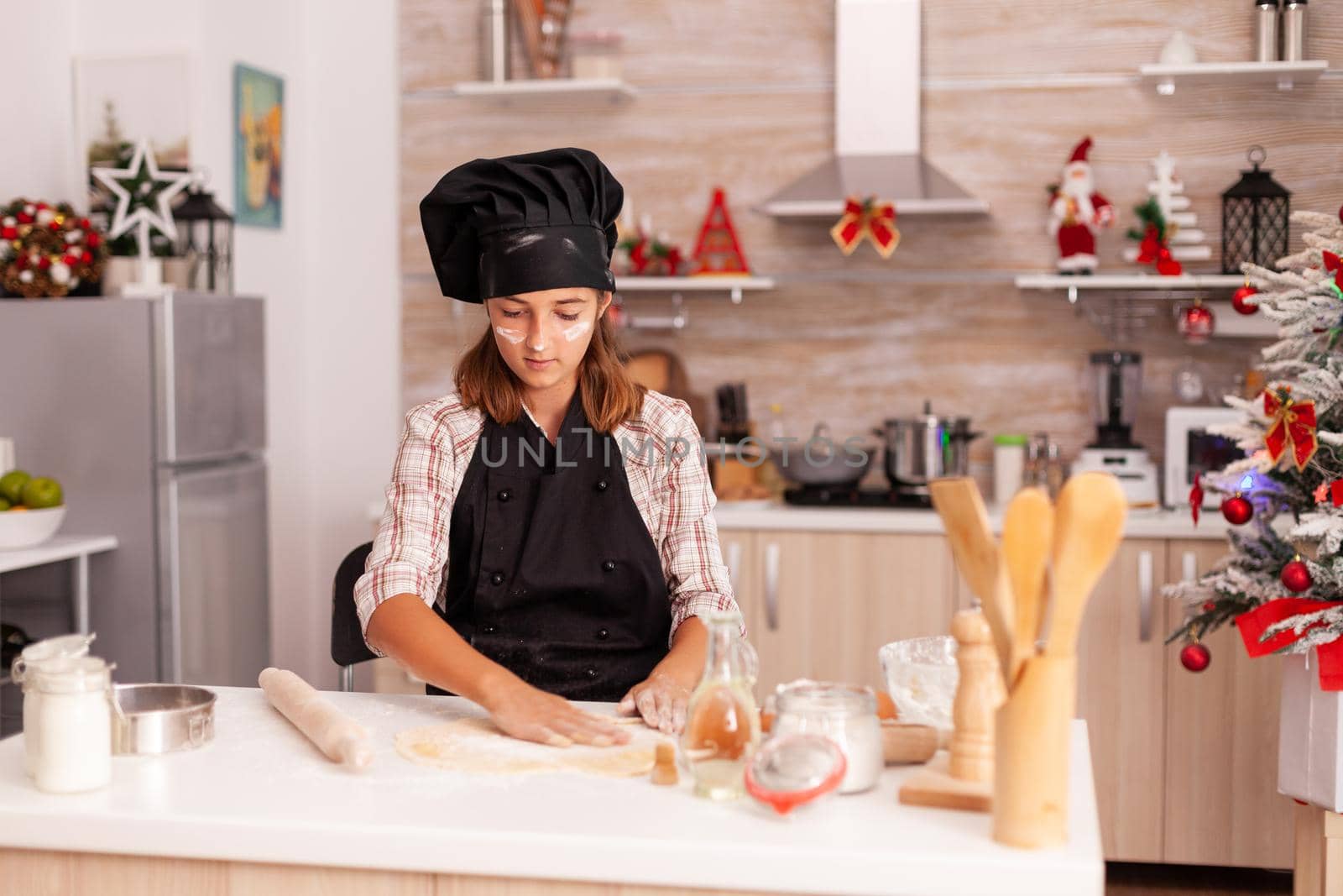 Child wearing baking apron putting flour on homemade dough preparing traditional gingerbread dessert enjoying winter holiday in xmas decorated kitchen. Kid celebrating christmas season making cookies