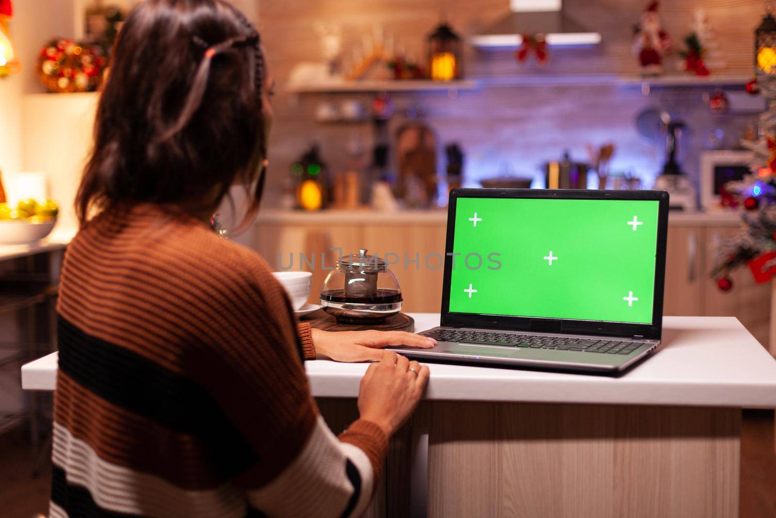 Modern young adult watching green screen on laptop digital device sitting at festive counter kitchen. Woman using computer chroma key gadget on background display for mockup template