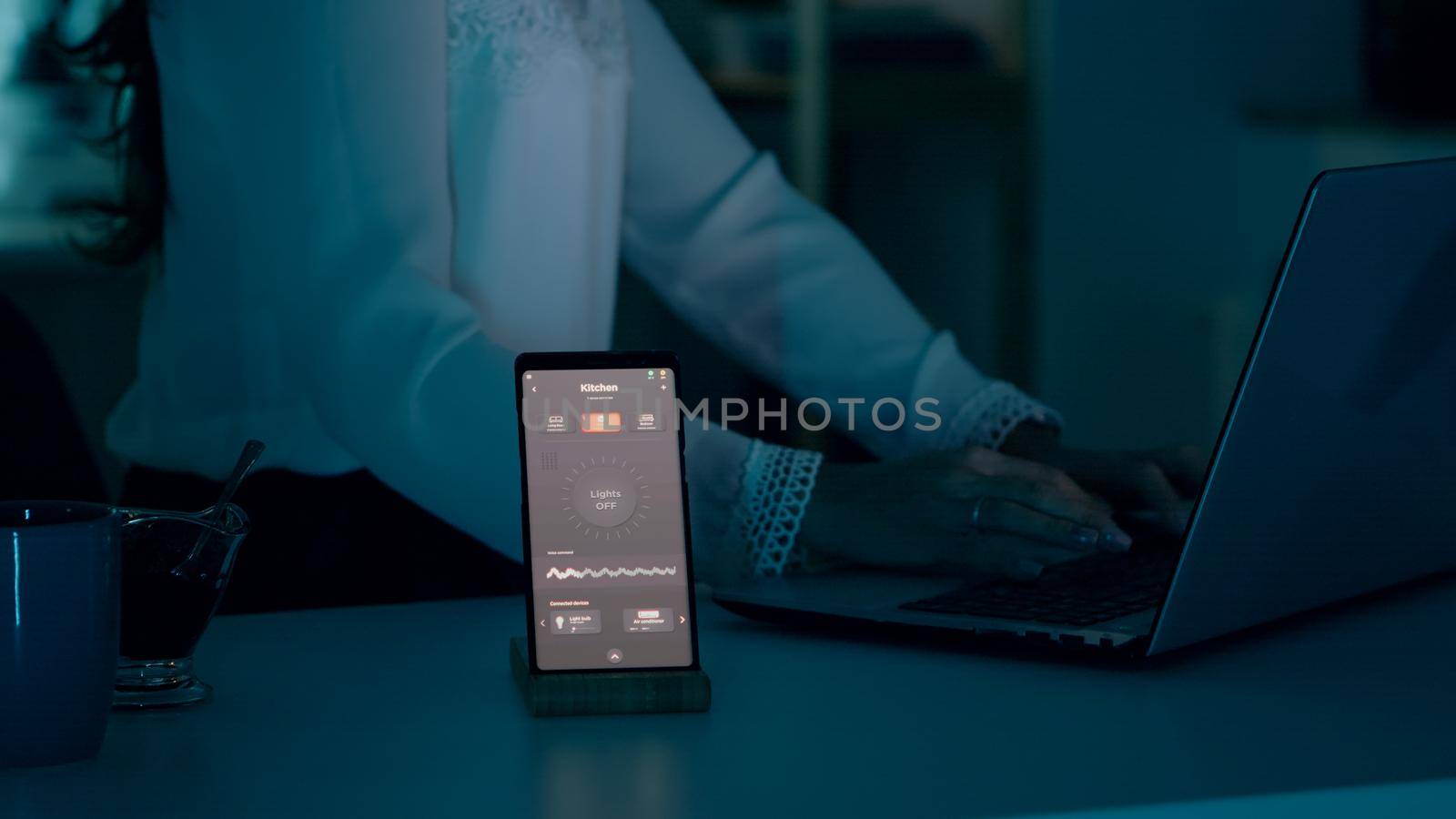 Woman typing at laptop sitting in home with automation lighting system using voice controlled on smartphone turning on bulbs. Speaker gadget responds to commands, person controlling ambiance light