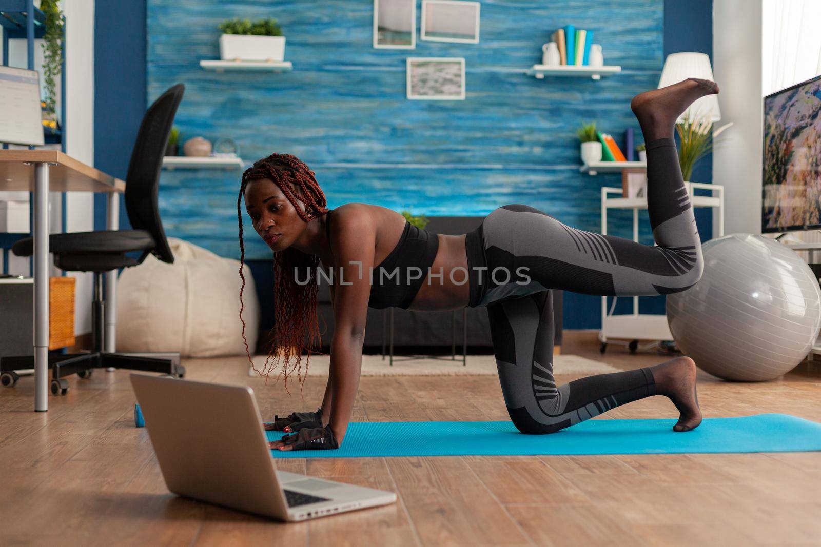 Black woman doing straight- leg donky kick pushing left foot up keeping leg bent, watching tutorial video on laptop screen, wearing sportwear on yoga mat for muscle strenght andn healthy lifestyle.