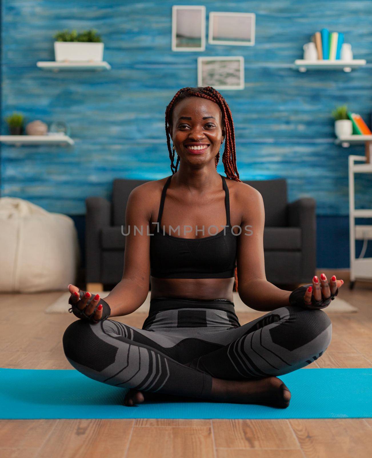 Portrait of black woman smiling practicing yoga at home sitting in lotus pose on mat mediting, practicing mindfulness meditation harmony, African american person doing relaxing workout.