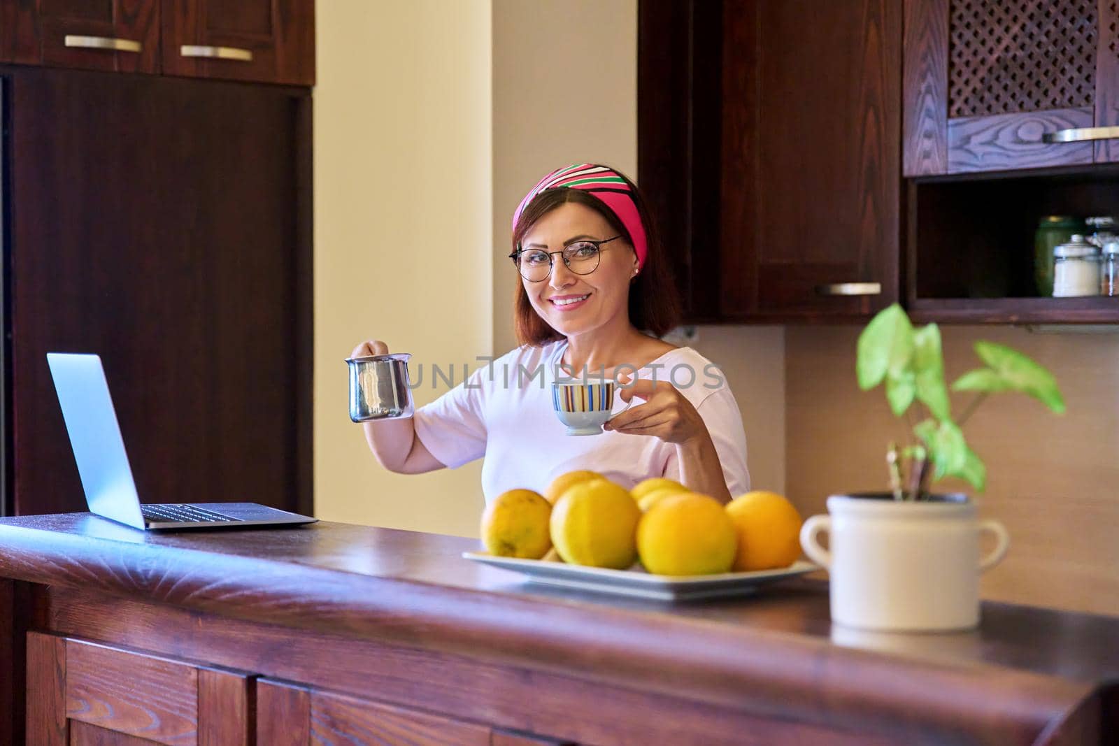 Smiling relaxing woman with morning coffee with cezve turk and laptop in the kitchen at home. Middle-aged female enjoying a drink, technology, lifestyle, home