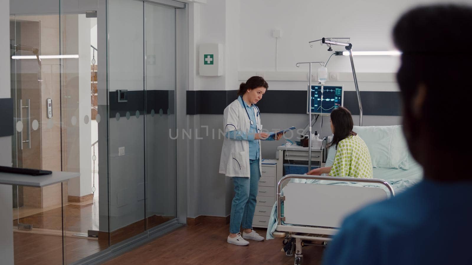 Medical team checking patient vital monitoring heart rate helping with fluids drip bag for remission therapy in hospital ward. Sick woman waiting for healthcare treatment during clinic examination