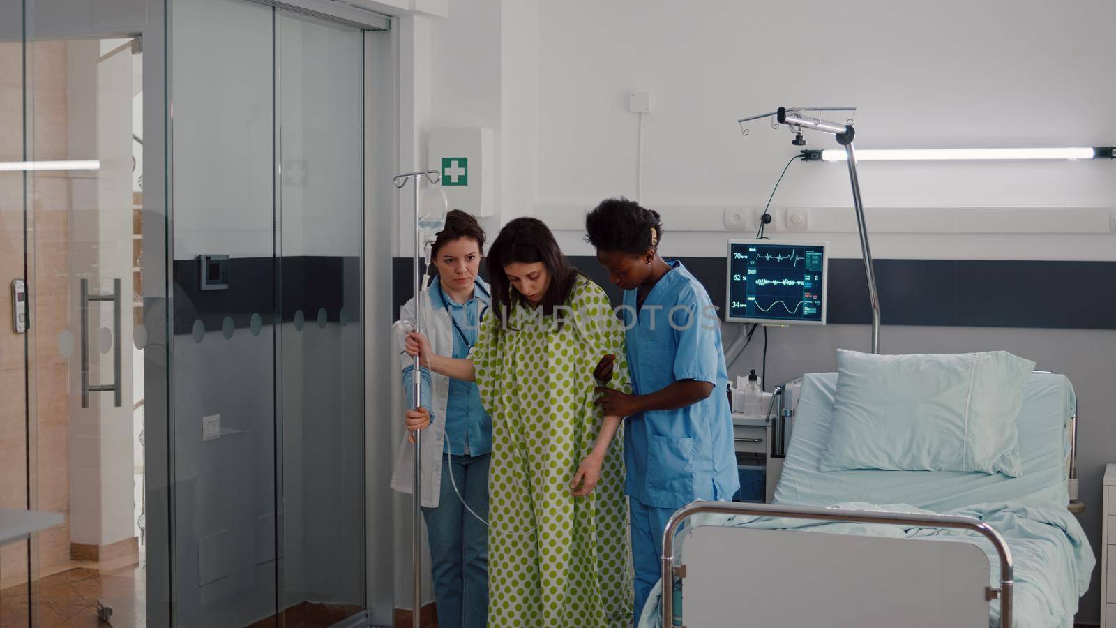 Afro american nurse with doctor helping patient to stand up from bed holding intravenous IV fluid drip bag after medical surgery in hospital ward. Practitioner monitoring sickness recovery