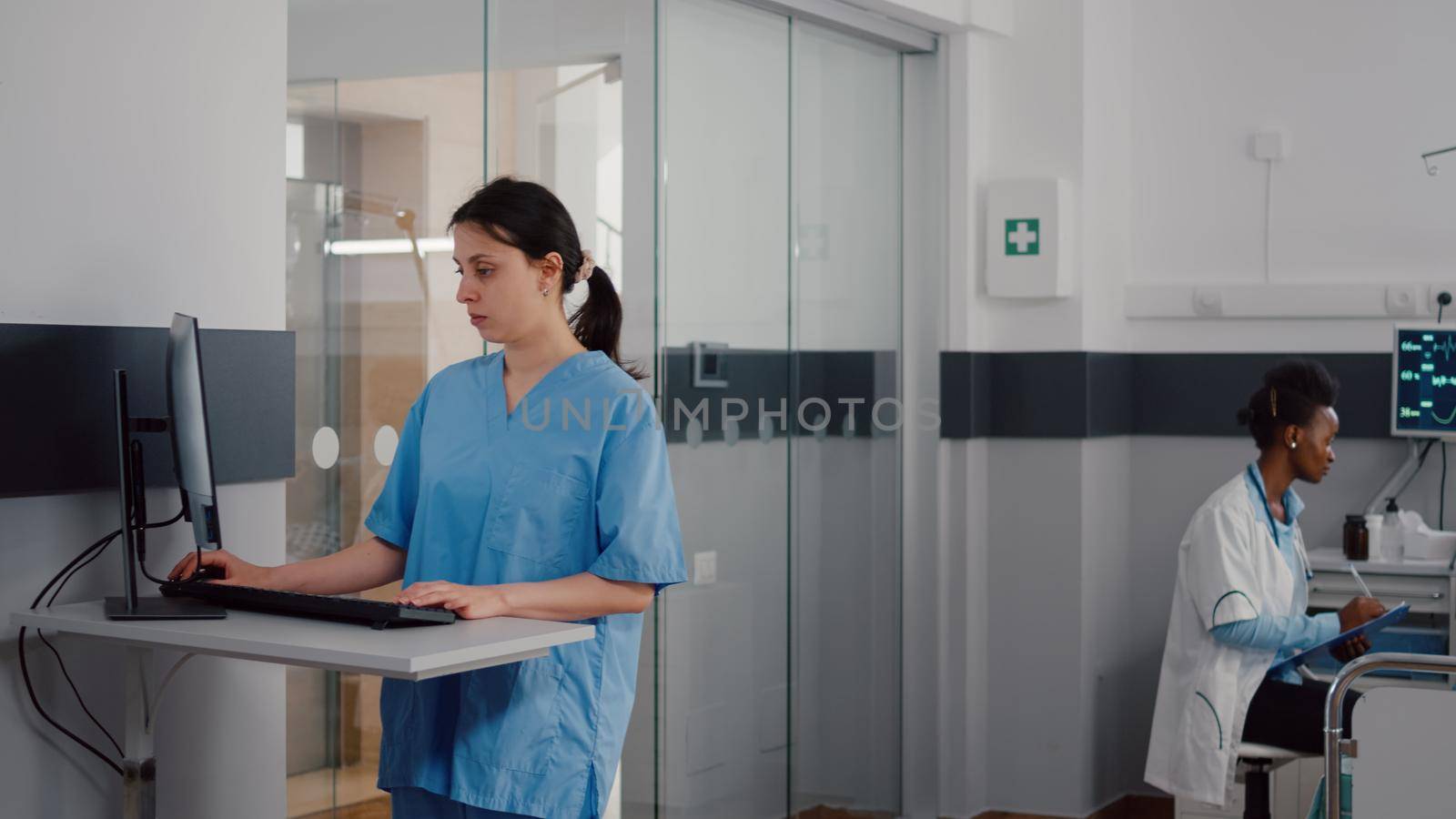 Front view of assistant typing medical expertise on computer while in background afro american practitioner talking with sick man healthcare treatment. Hospitalized patient having respiratory disorder