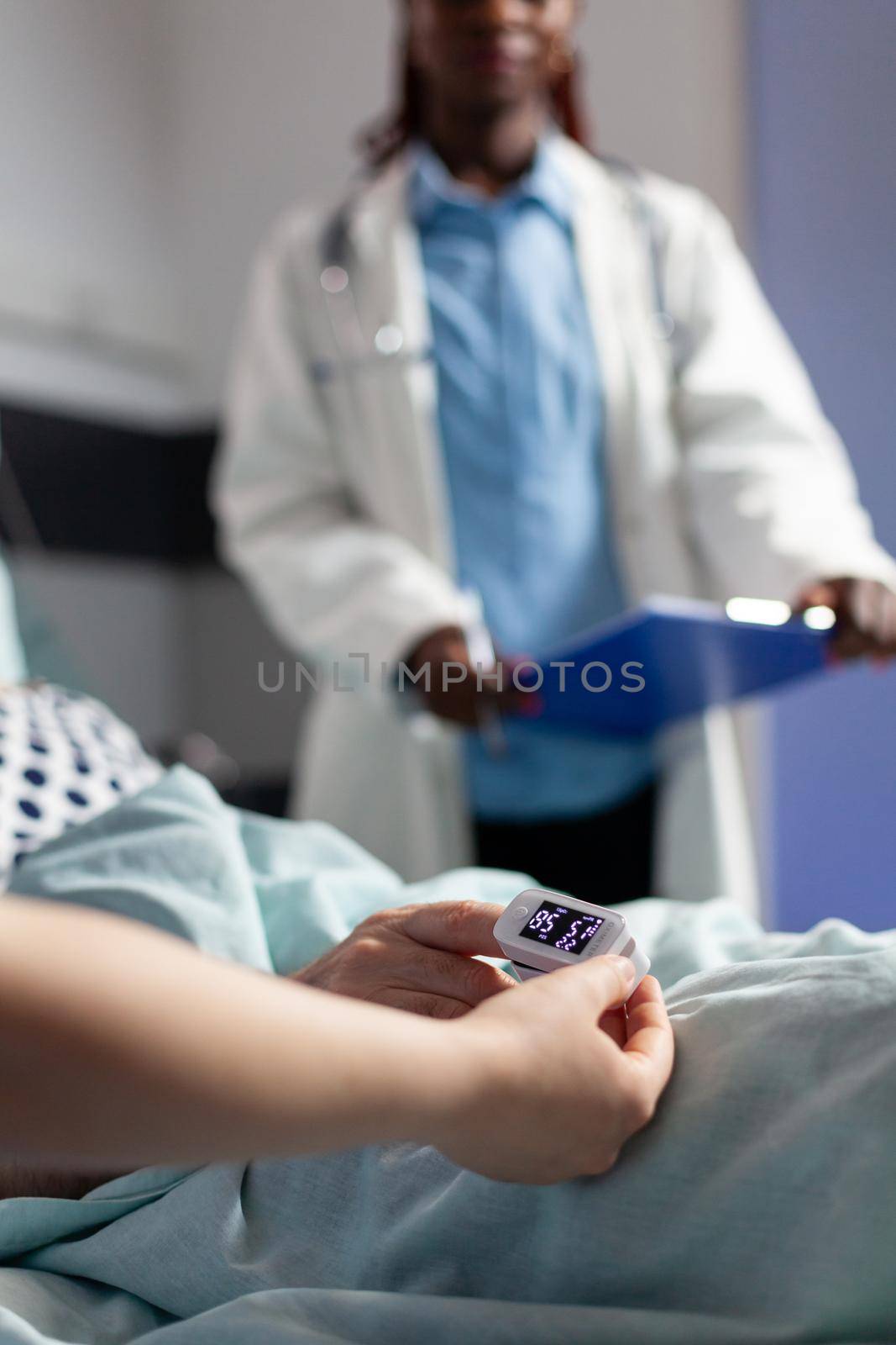 Close up of oxymeter attached on sick senior patient laying in hospital bed, monitoring hemoglobin. Medical staff using modern technology for reading blood oxygen saturation.