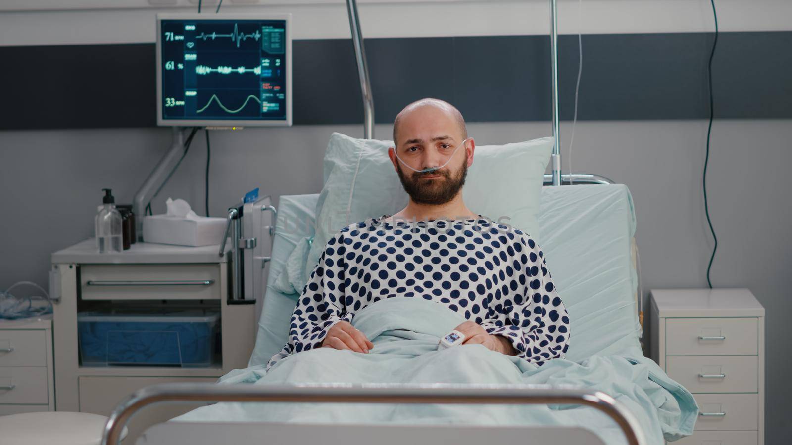 Potrait of sad sick man wearing nasal oxygen tube lying in bad looking into camera during sickness recovering in hospital ward. Depressed patient waiting for respiratory treatment