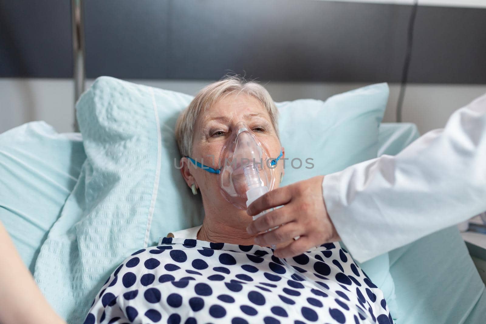 Doctor and nurse supervising senior woman breathing with oxygen mask, laying in bed because of pulmonary disease. Patient laying in bed with iv dip bag attached.