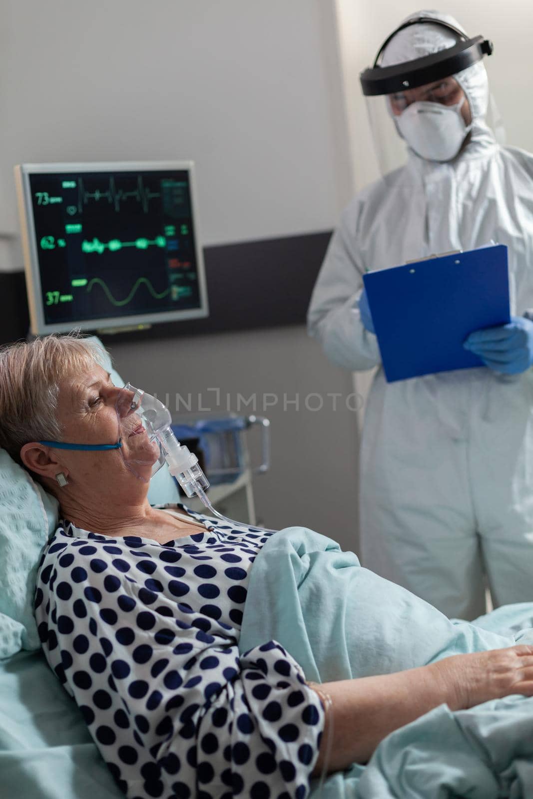 Sick senior woman inhale and exhale through oxygen mask laying in hospital bed during global pandemic with coronavirus. Doctor dressed in coverall pee suit to prevent infection with covid-19.